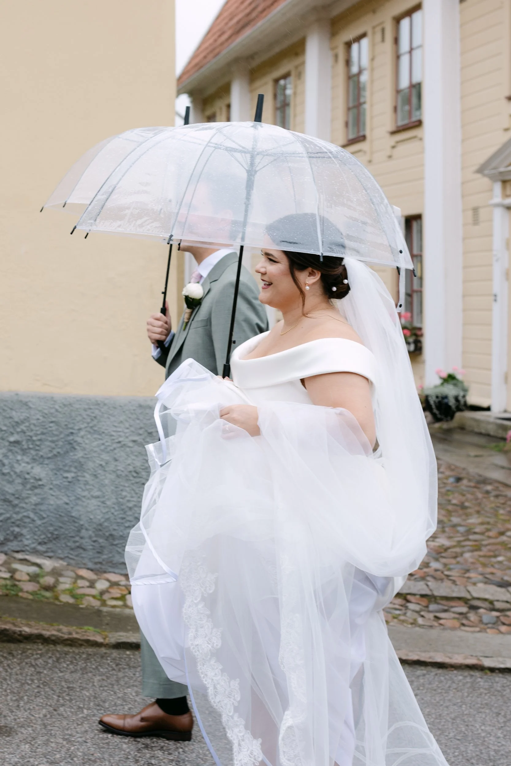 En glad brud i vit brudklänning går utomhus under ett transparent paraply, med en man i grå kostym och bruna skor, framför en gul och beige byggnad på en grå gatsten.