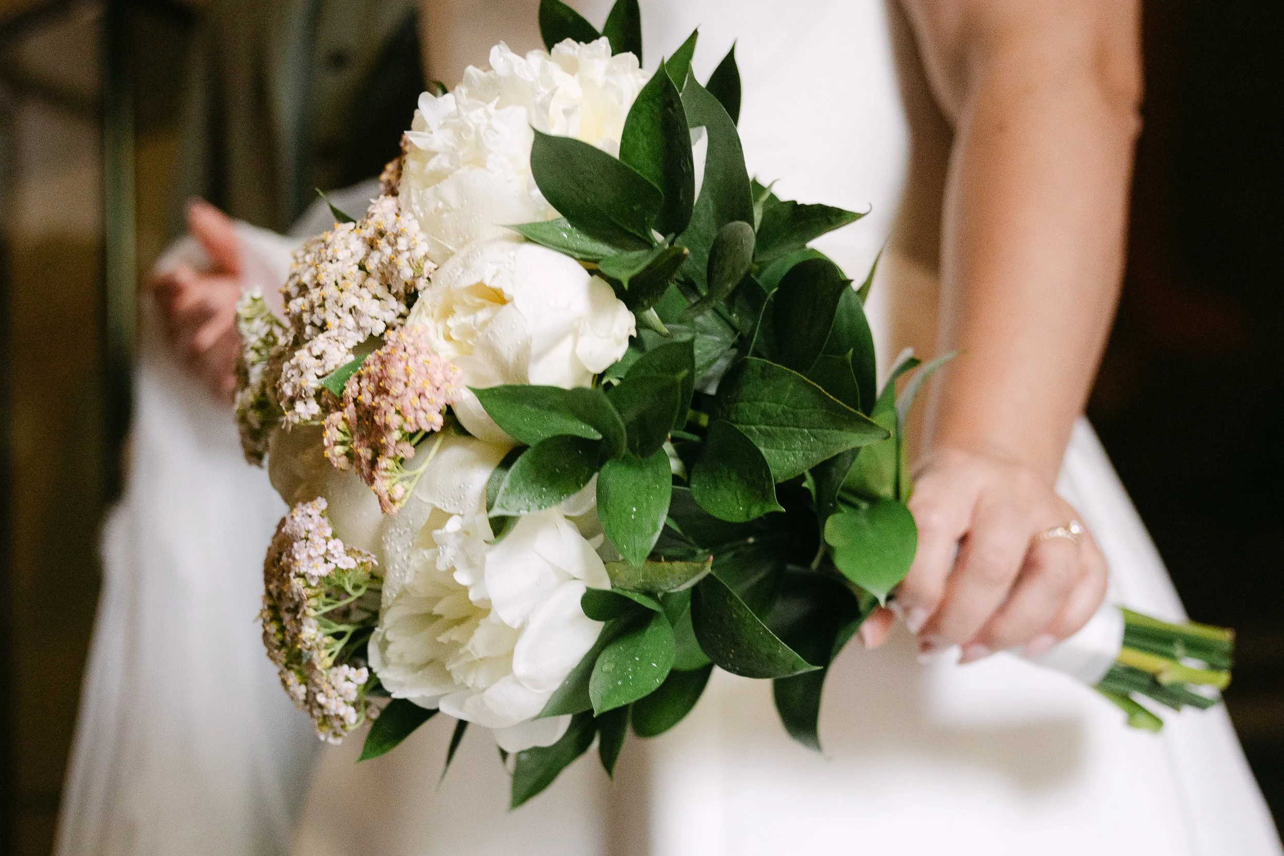En person håller i en bukett med vita blommor och gröna blad, antagligen en bröllopsbukett.
