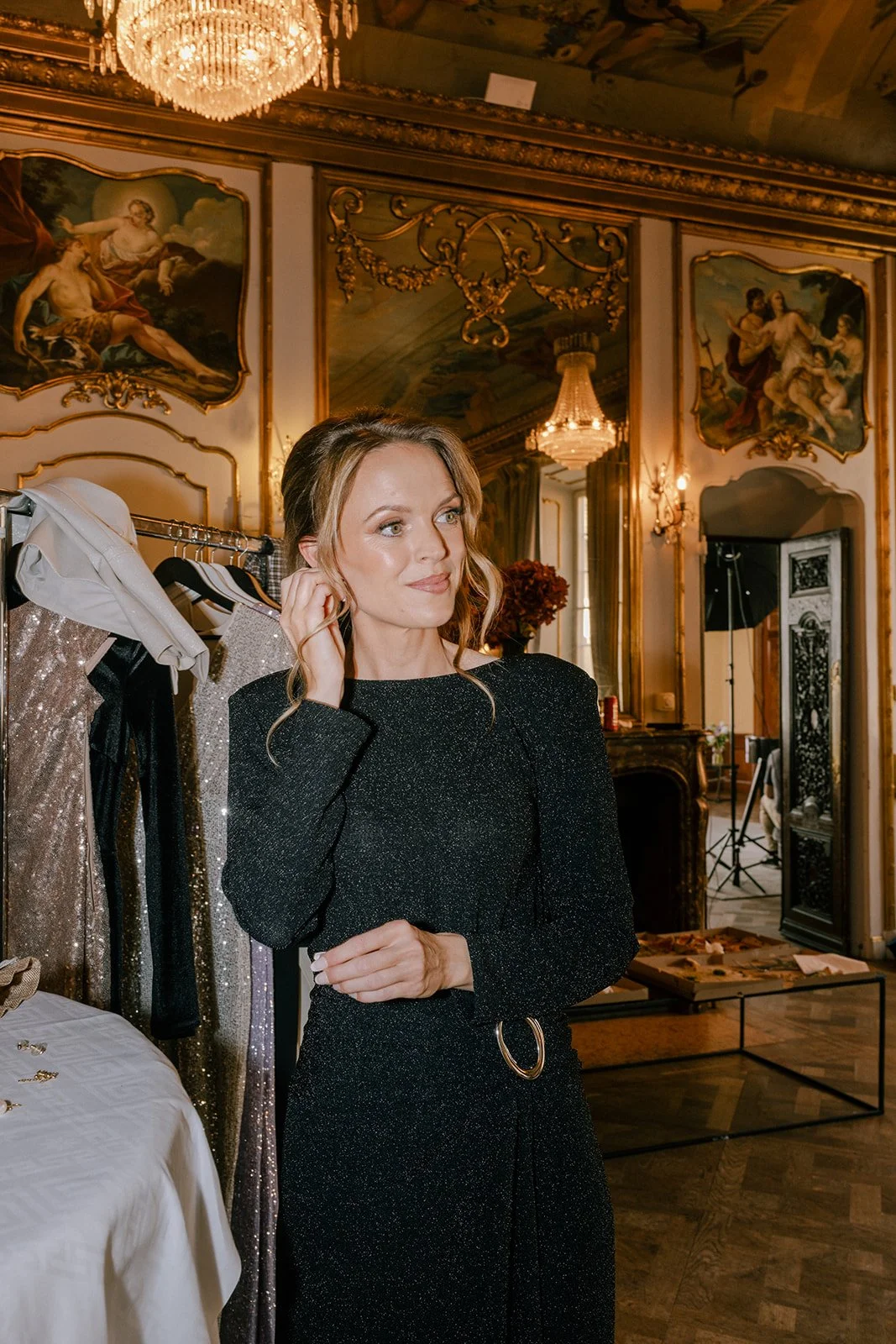 En kvinna står i ett elegant rum med konst och takmålningar, iklädd en svart klänning med glitter. Hon justerar håret nära örat.
