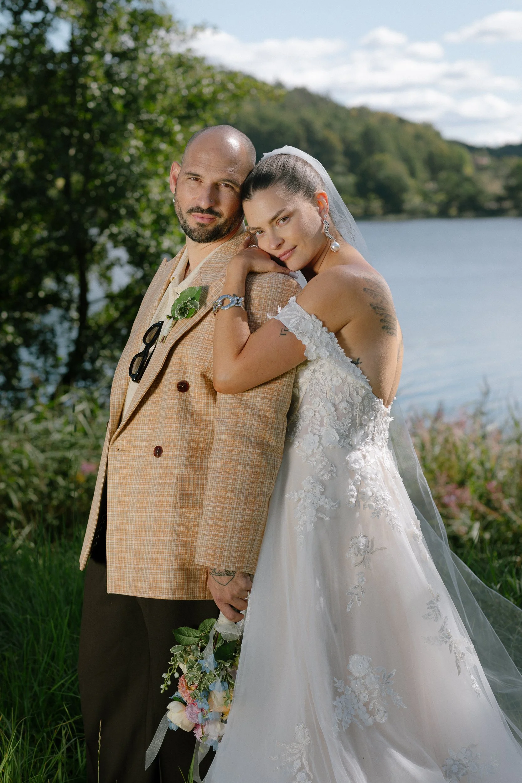 En nygift man och kvinna poserar utomhus vid en sjö, kvinnan bär bröllopsklänning och mannen en beige blazer, med grön natur i bakgrunden.  Av Sofia Wilhelmina bröllopsfotograf i Stockholm.