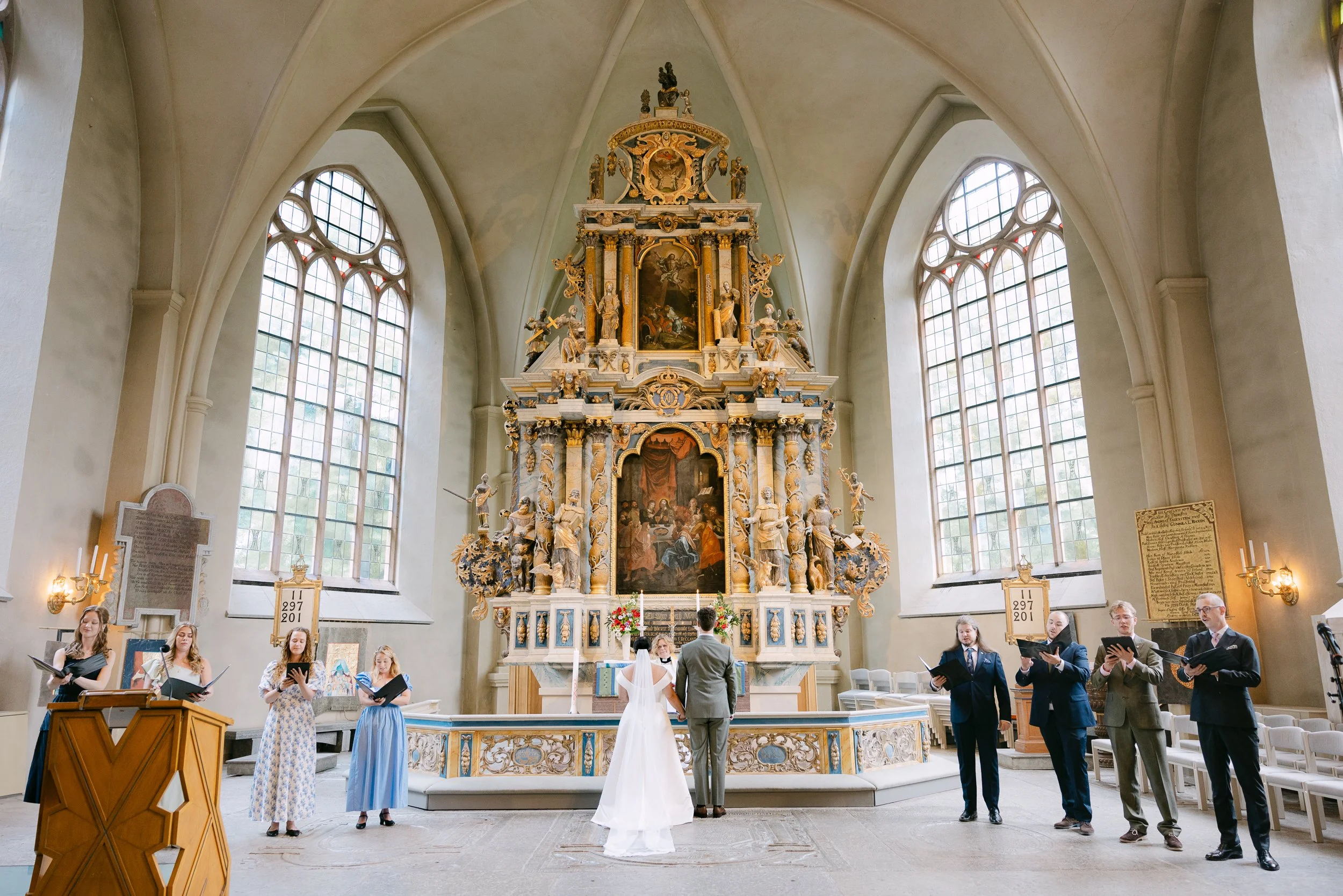 Firetonad kyrka med ett stort altare och fönster på vardera sida. En brud och en gäst i bröllopskläder står framför altaret, medan andra personer är samlade runt. Några personer sjunger eller reciterar ur ett psalmbok.