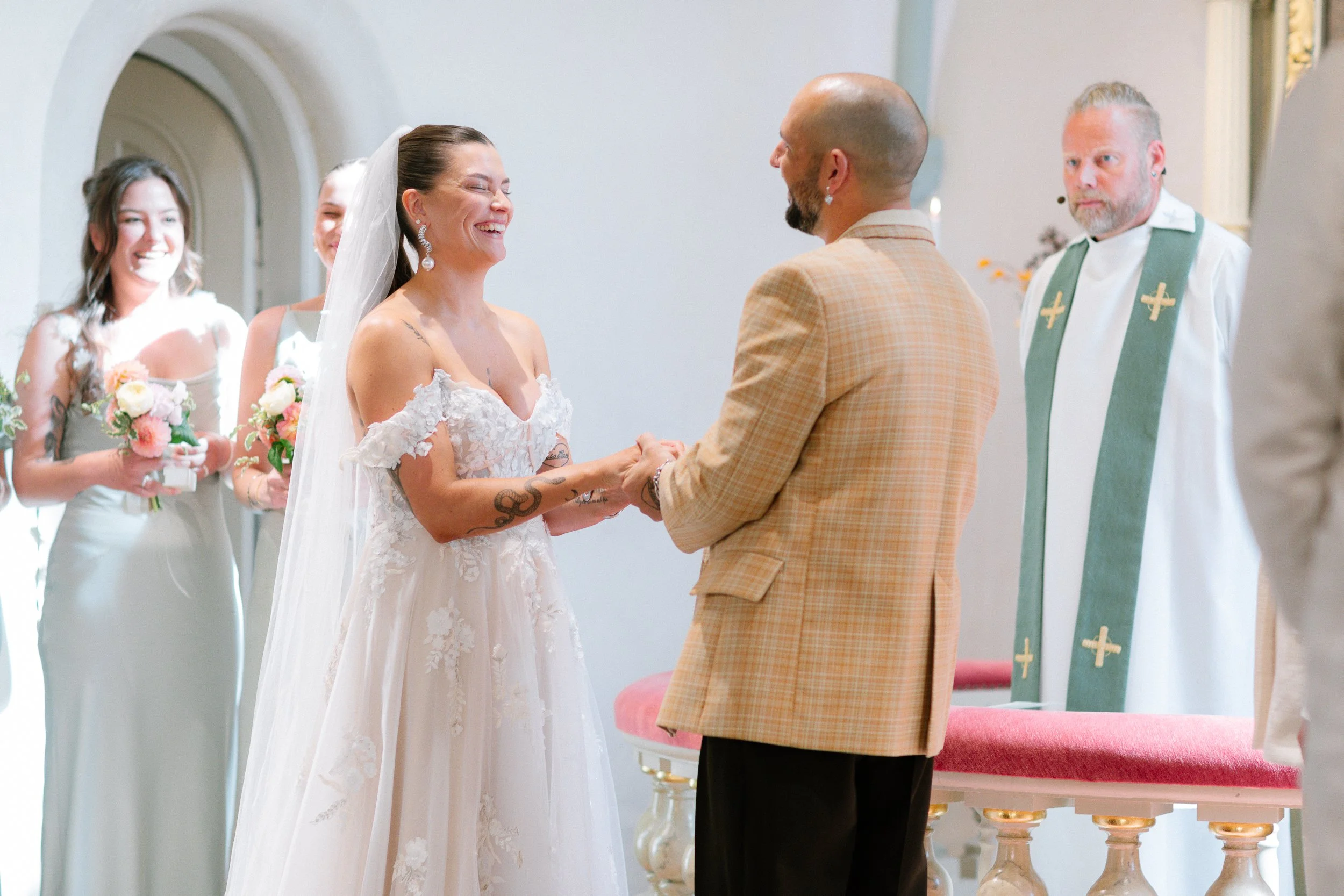 En brud och en brudgum håller hand under ett bröllopsceremoni i en kyrka, omgiven av brudtärnor och en präst.  Av Sofia Wilhelmina bröllopsfotograf i Stockholm.