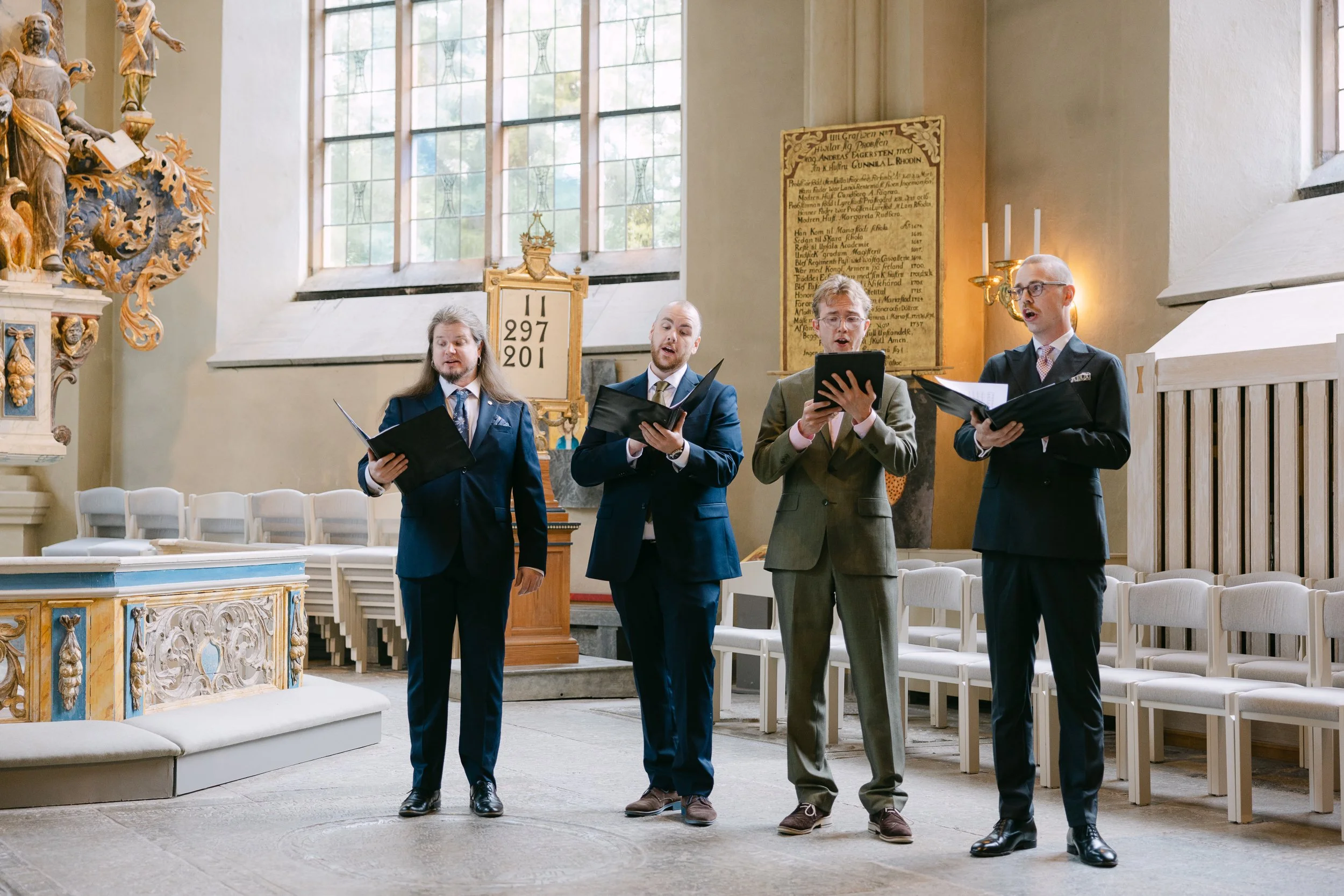 Fyra män i mörka kostymer sjunger i en kyrka, med handhållen notblad, i ett lugnt och elegant inomhusmiljö med stora fönster och utsmyckad altar.