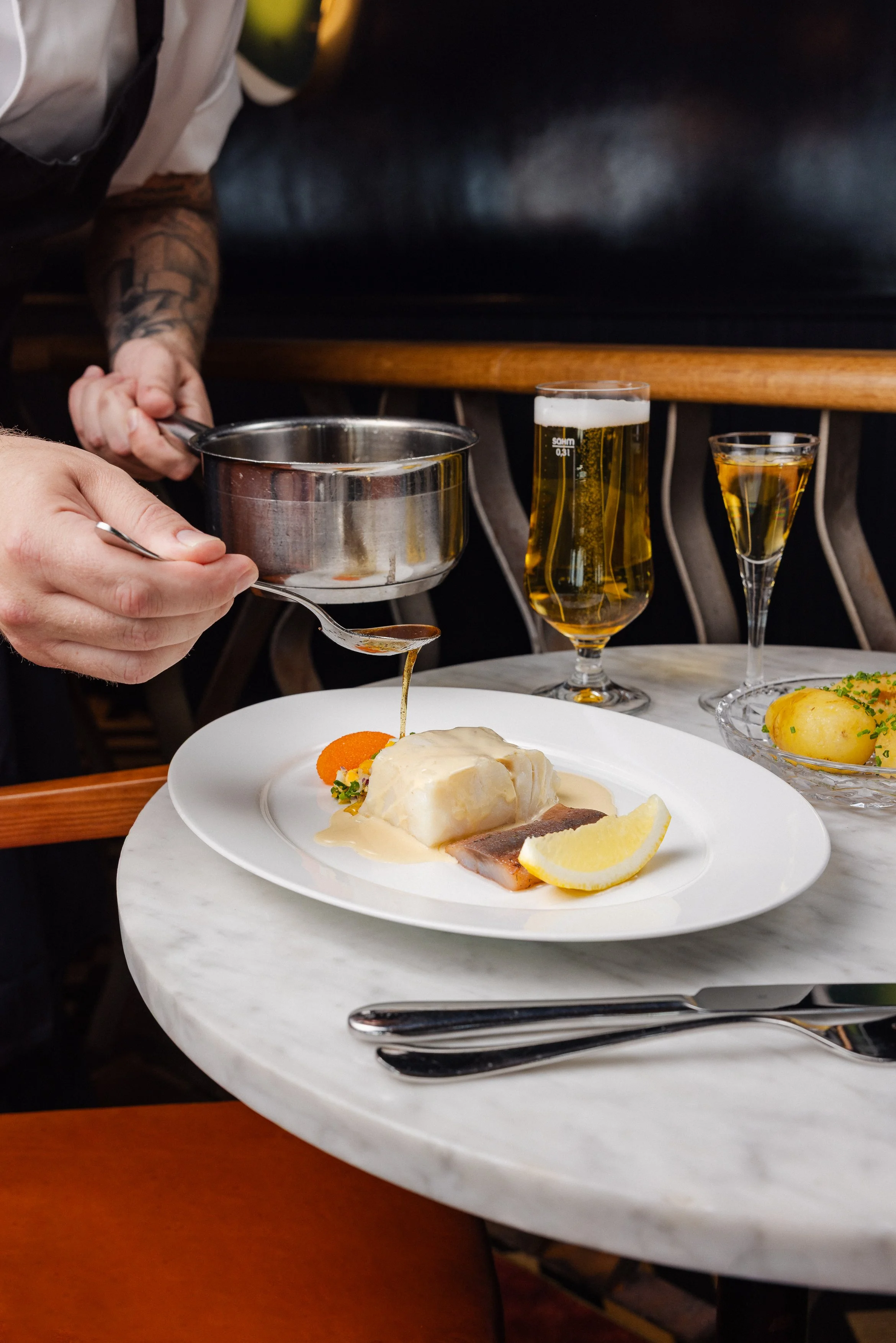 En person som häller sås över en fiskrätt på ett vitt serveringsfat, med en citronklyfta, ett kokt ägg, en morot och grönsaker, på ett runt marblebord, med öl och en drink i bakgrunden. Reklamfoto, restaurang, Gondolen Stockholm. 
