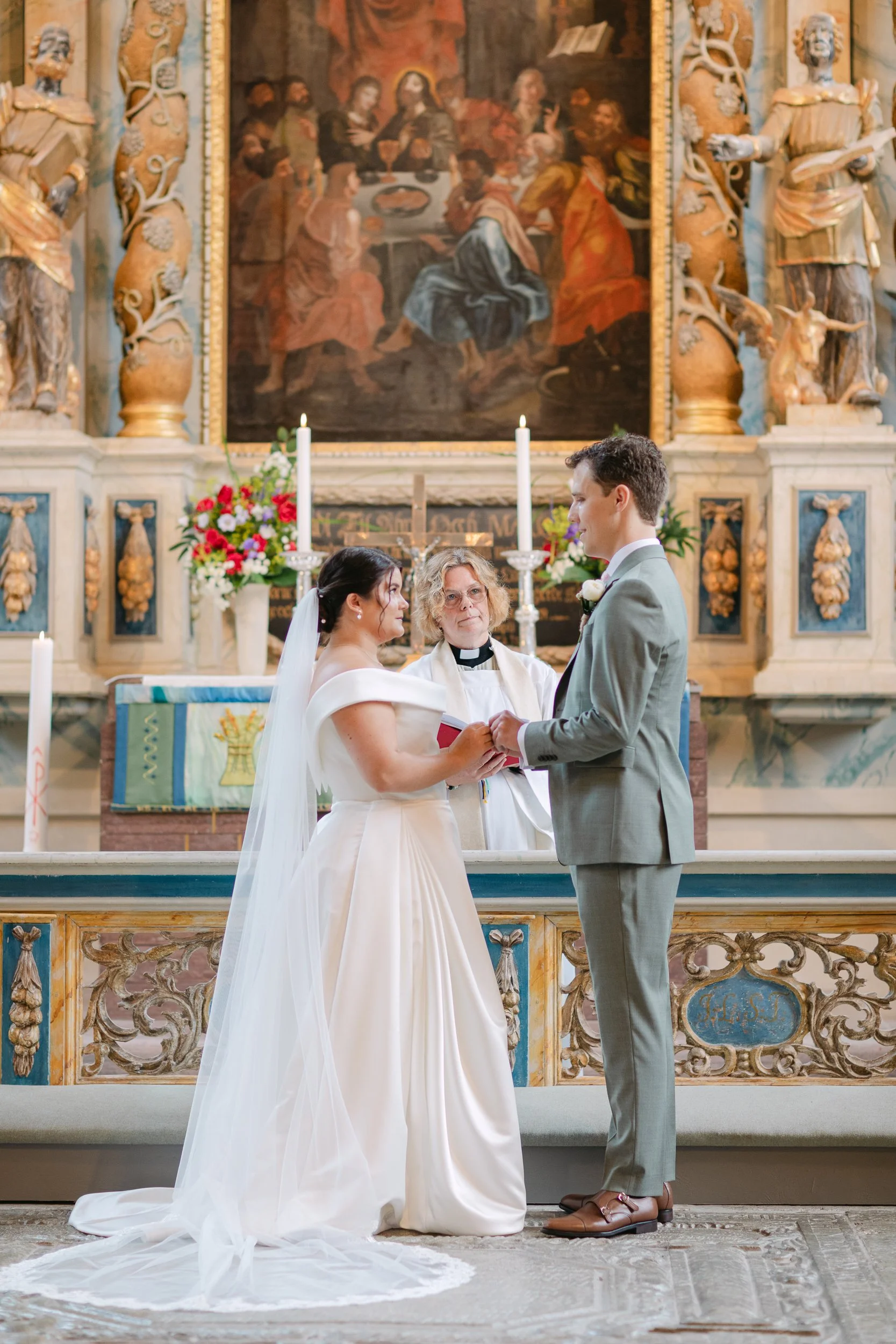En brud och en brudgumen håller hand under ett bröllopfirande i en kyrka. Ekumenisk präst står mellan dem. Bakom dem finns altaret dekorerat med blommor och ljus, och en stor religiös målning.