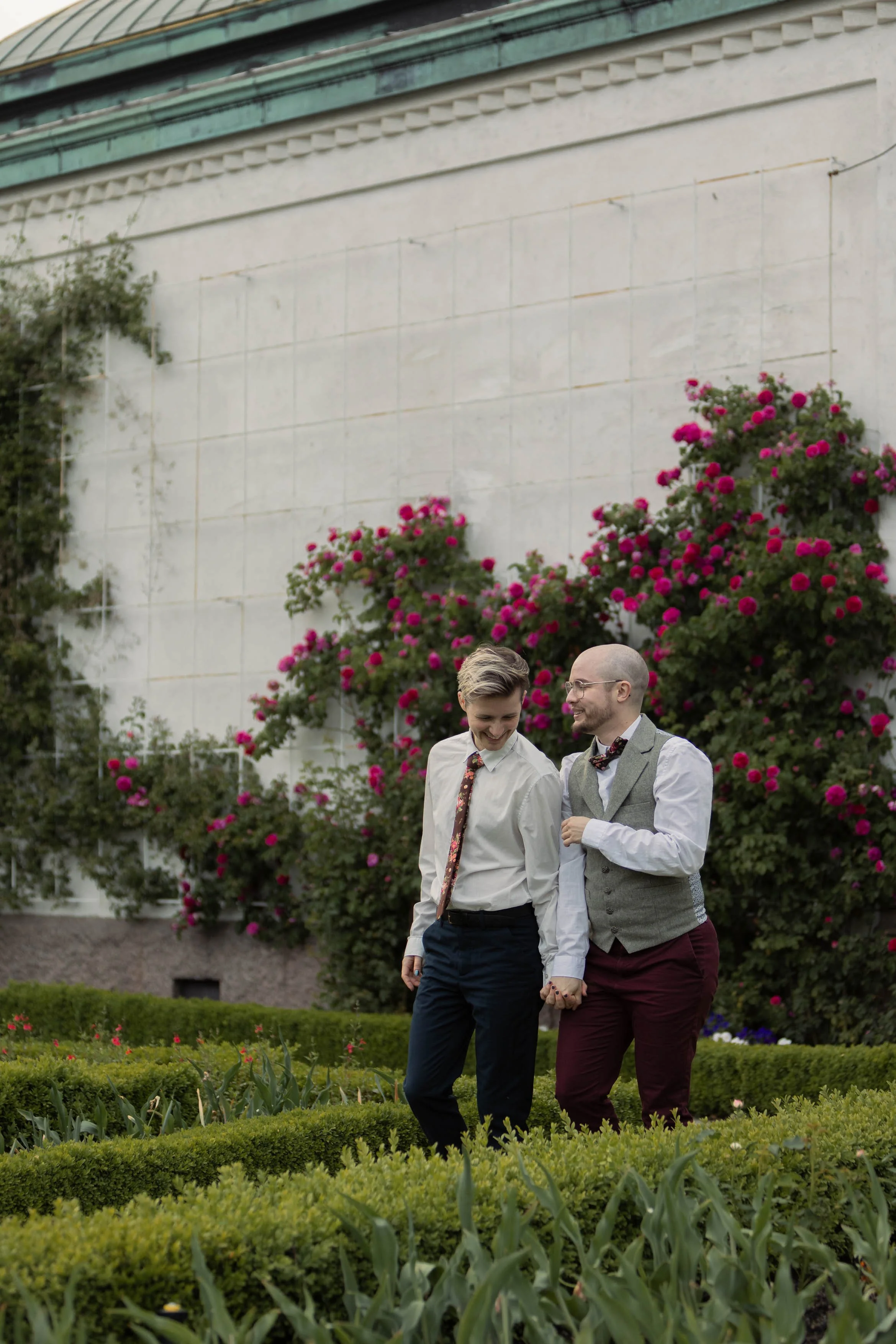 Två personer i formella kläder håller varandra i handen och går i en trädgård med rosa blommor och gröna buskar, en vägg med rosa blommor i bakgrunden.