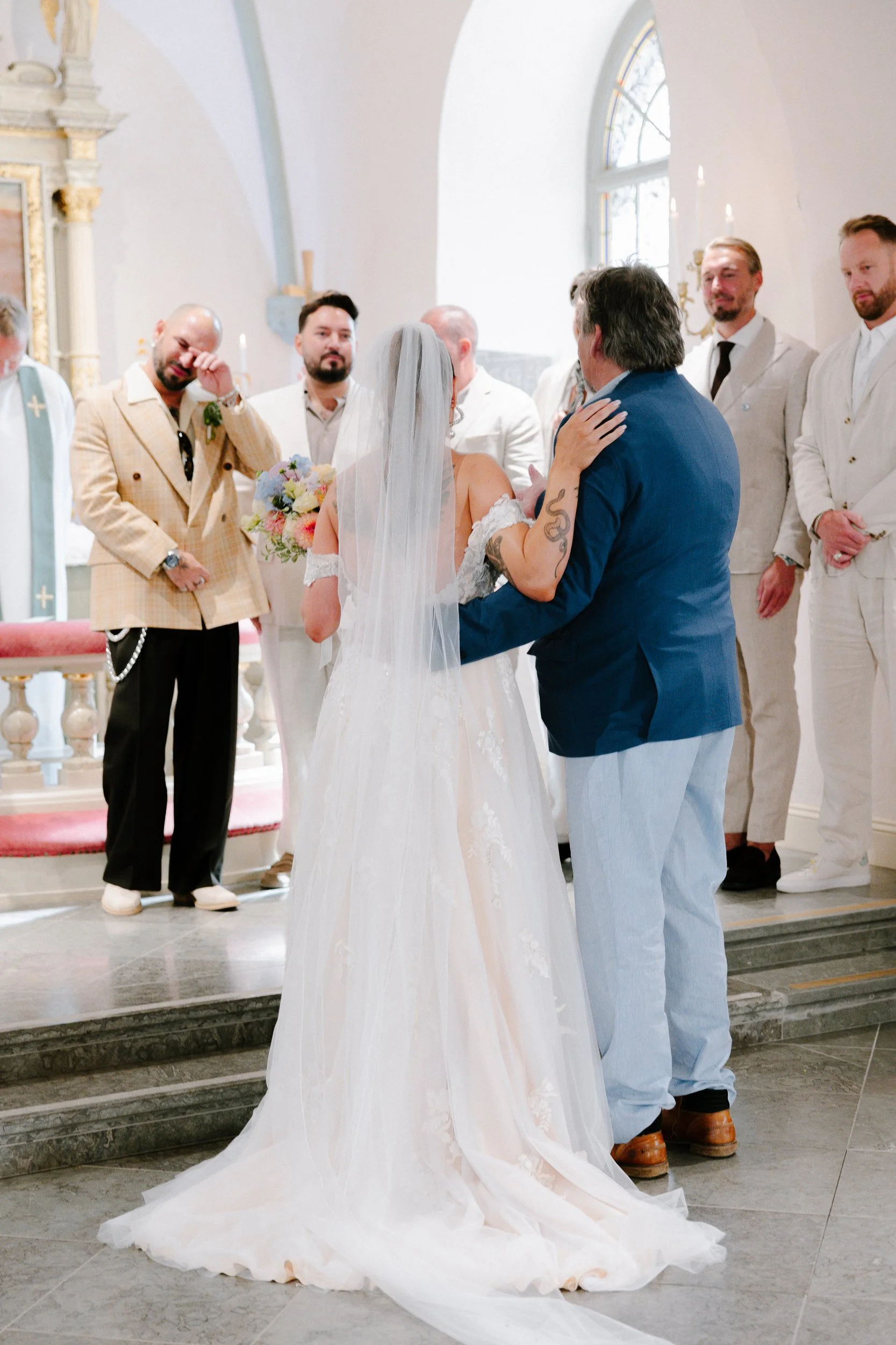 En brud i bröllopsklänning och en man i blå kavaj håller tal eller pratar vid ett bröllop i en kyrka, med gäster i bakgrunden.  Av Sofia Wilhelmina bröllopsfotograf i Stockholm.
