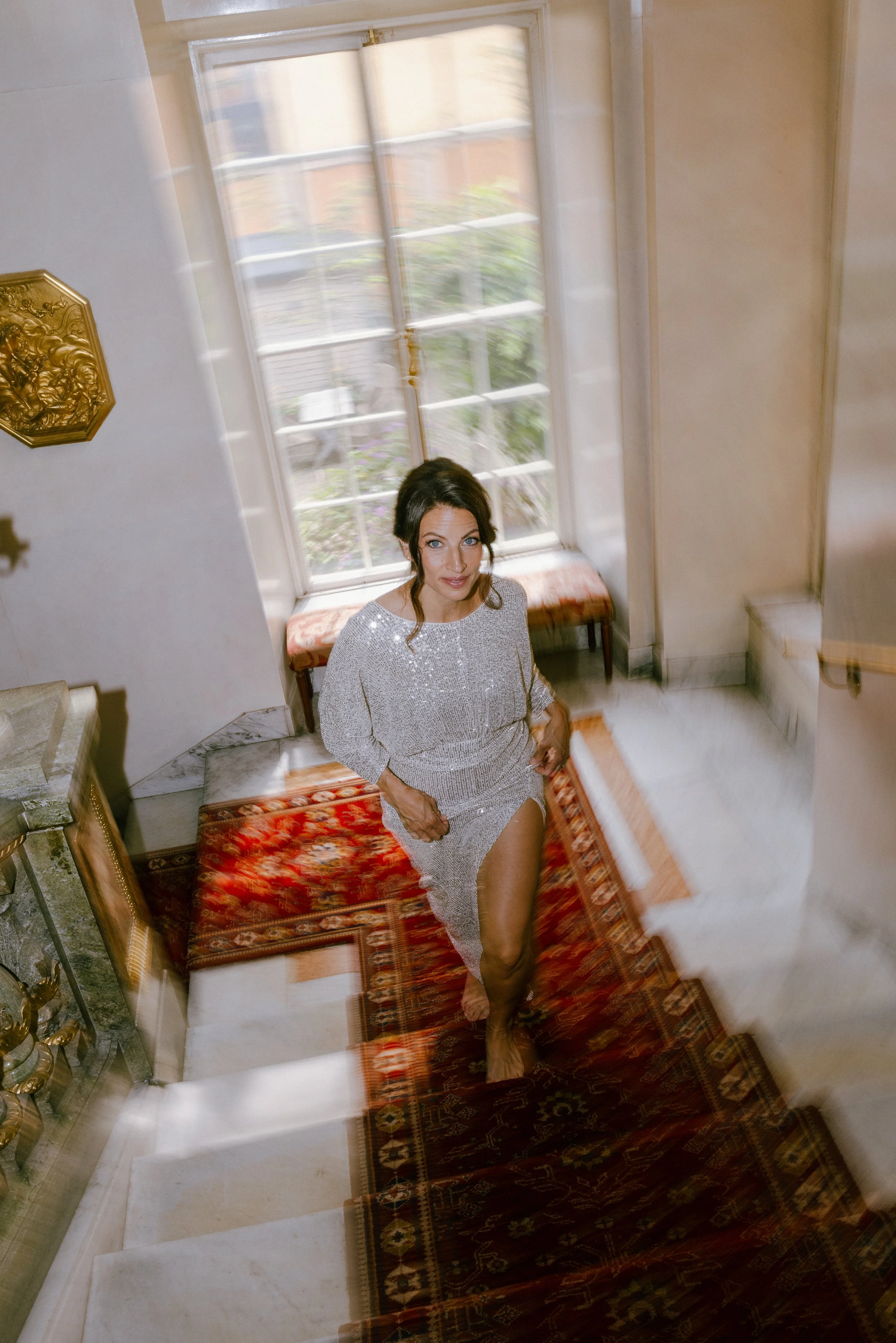 En kvinna i en glitterklänning står på en trappa inomhus, tittar upp mot kameran. Bakom henne finns ett stort fönster med utsikt mot en trädgård.