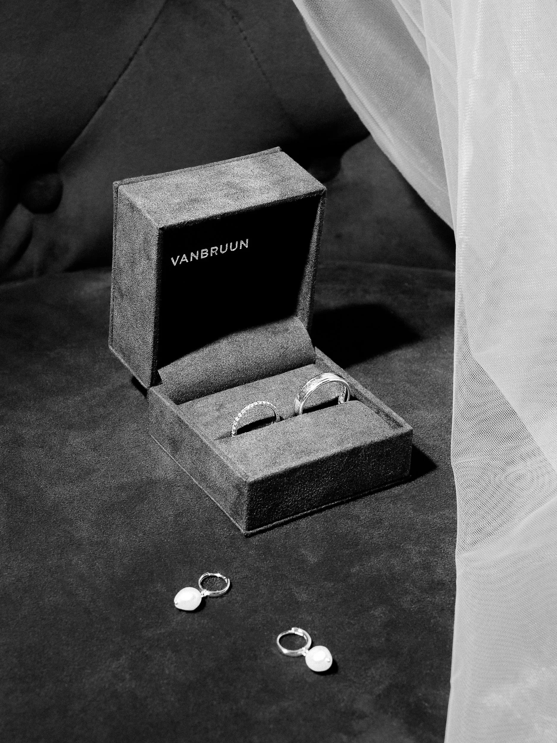 Svartvitt foto av ett presentbox med ringar, ett par ringar och ett par knappar på en svart yta. En person bär en ljus klar designad skjorta.