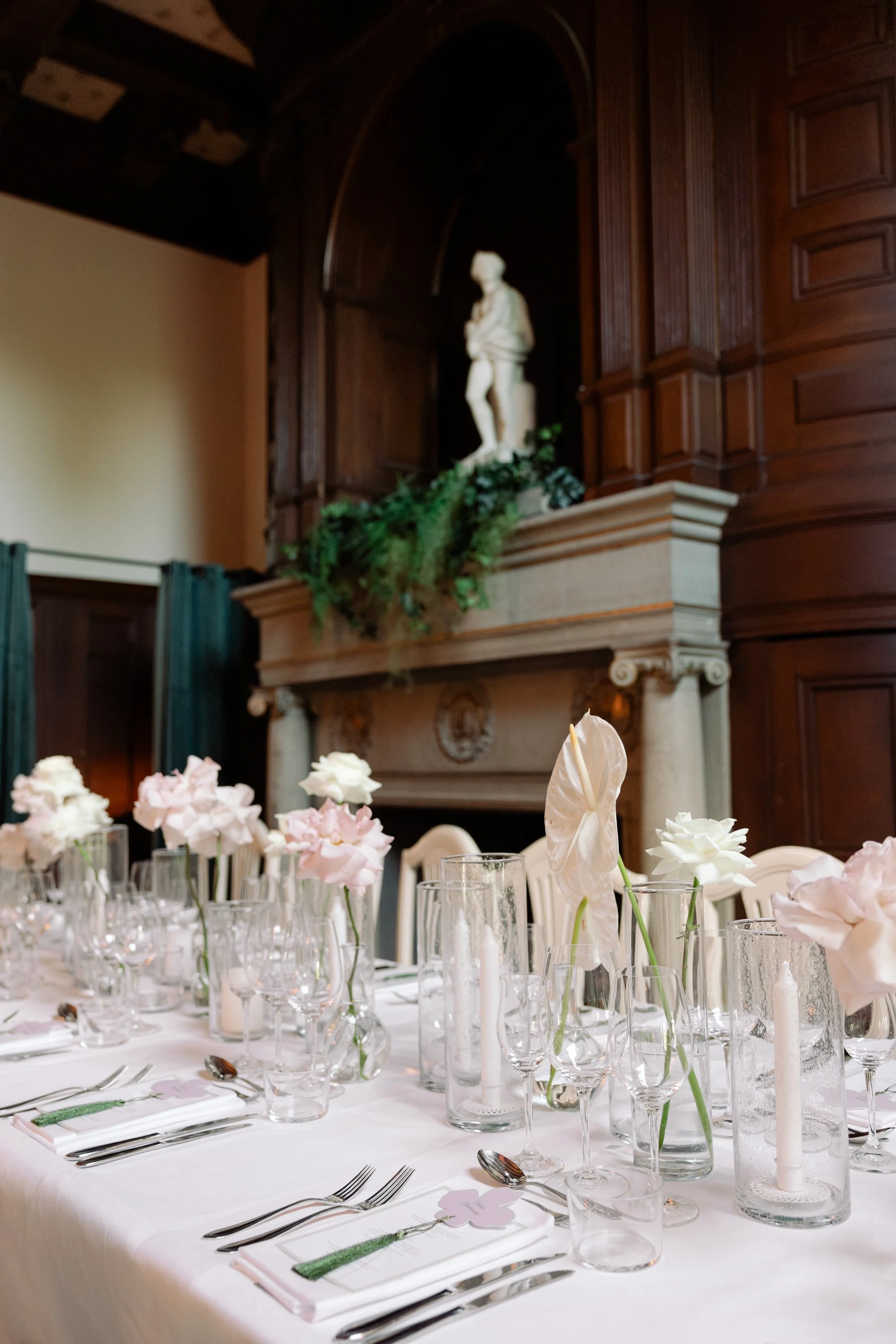 Vitt dukatbord med glas, bestick, blomsterarrangemang i glasvaser, och ljuslyktor i glas, i en elegant festlokal med träpanel och en skulptur på en piedestal i bakgrunden.