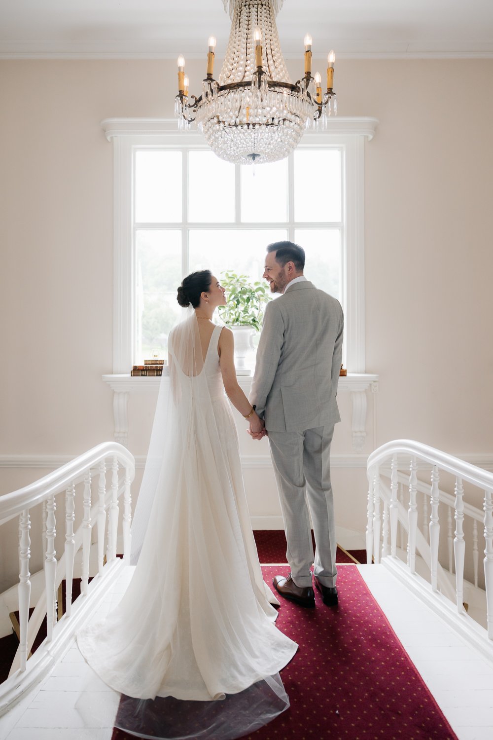 En nygift brud och brudgum står tillsammans på en trappa i ett ljust rum med stort fönster och kristallkrona, håller händer och tittar på varandra.