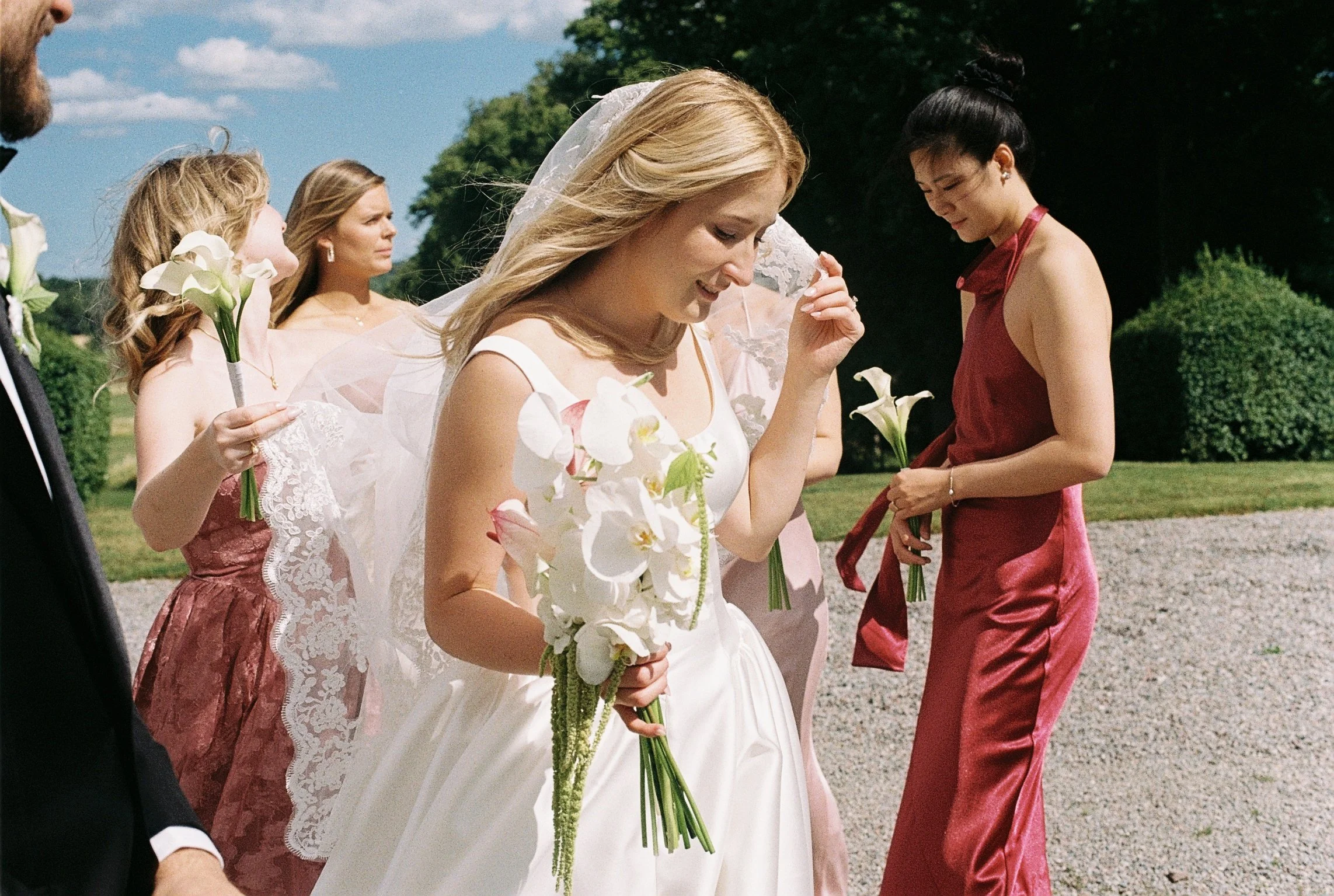 En grupp människor i formella kläder, inklusive en brud med bröllopsklänning och en brudnäbb med blommor, står utomhning under en klar himmel.