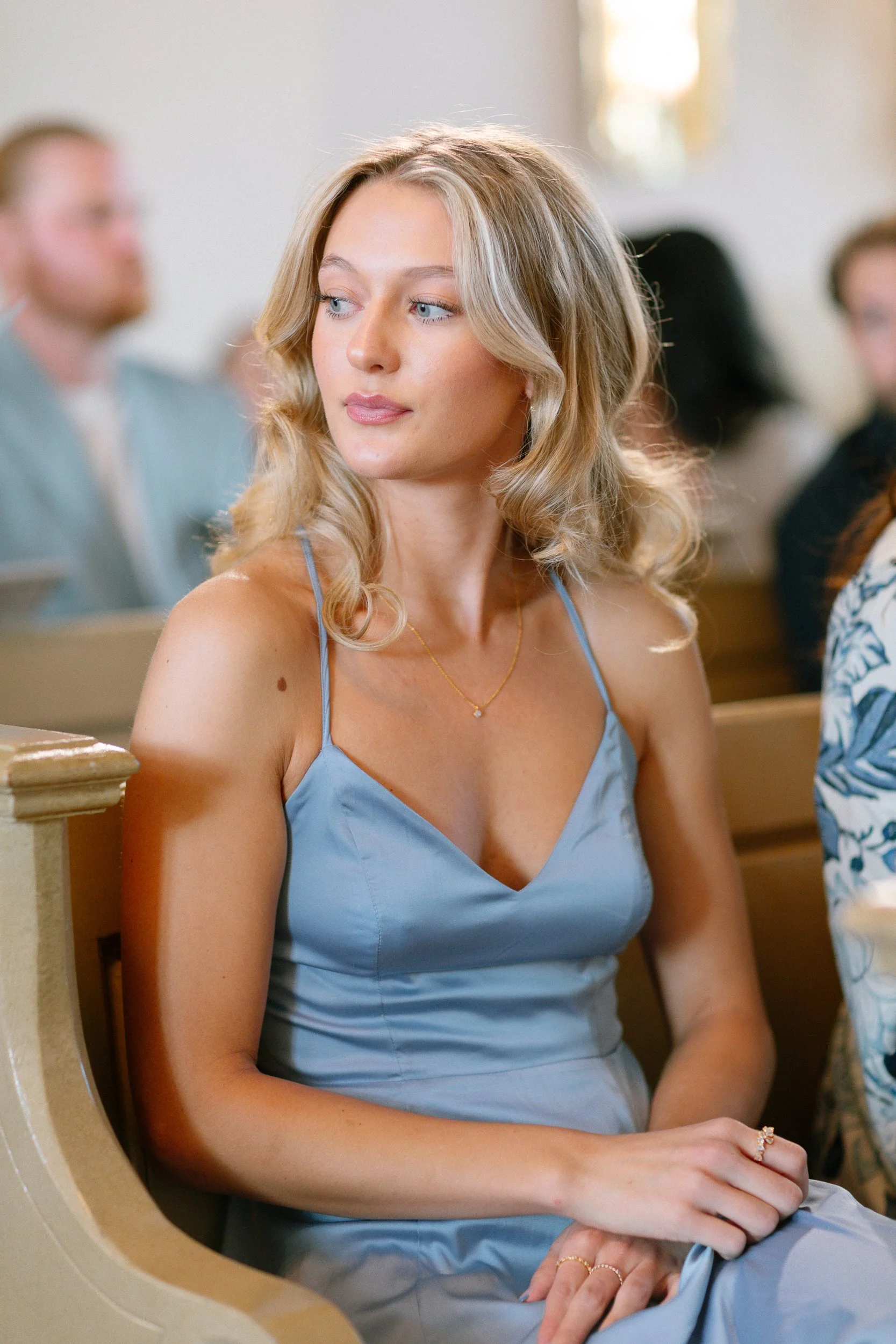 En ung kvinna med blont, lockigt hår bär en ljusblå klänning och sitter på bänk i en kyrka eller kapell, med andra personer i bakgrunden.  Av Sofia Wilhelmina bröllopsfotograf i Stockholm.