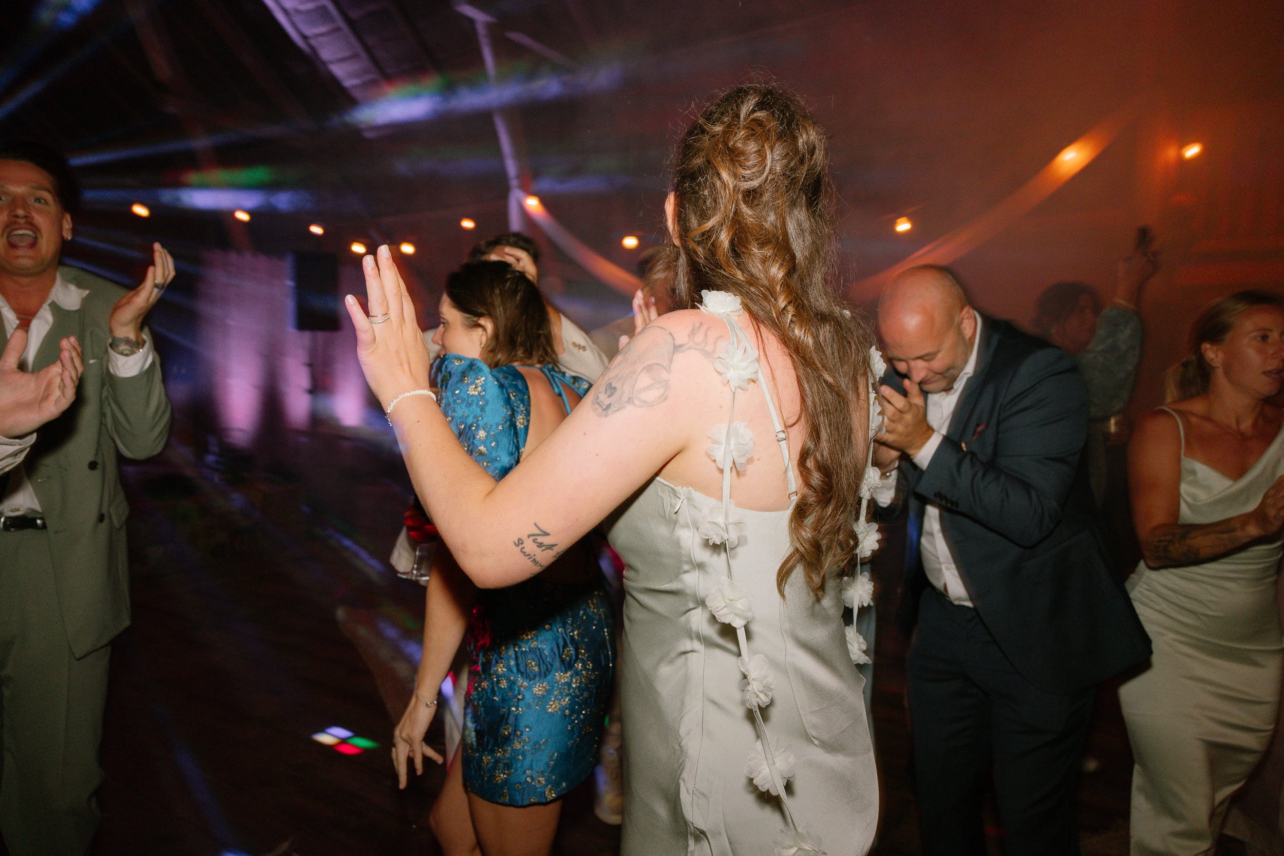 Folk dansar på en festlokal, vissa med händerna i luften och andra med huvudet sänkt, i en dimmig och ljusmiljö.  Av Sofia Wilhelmina bröllopsfotograf i Stockholm.