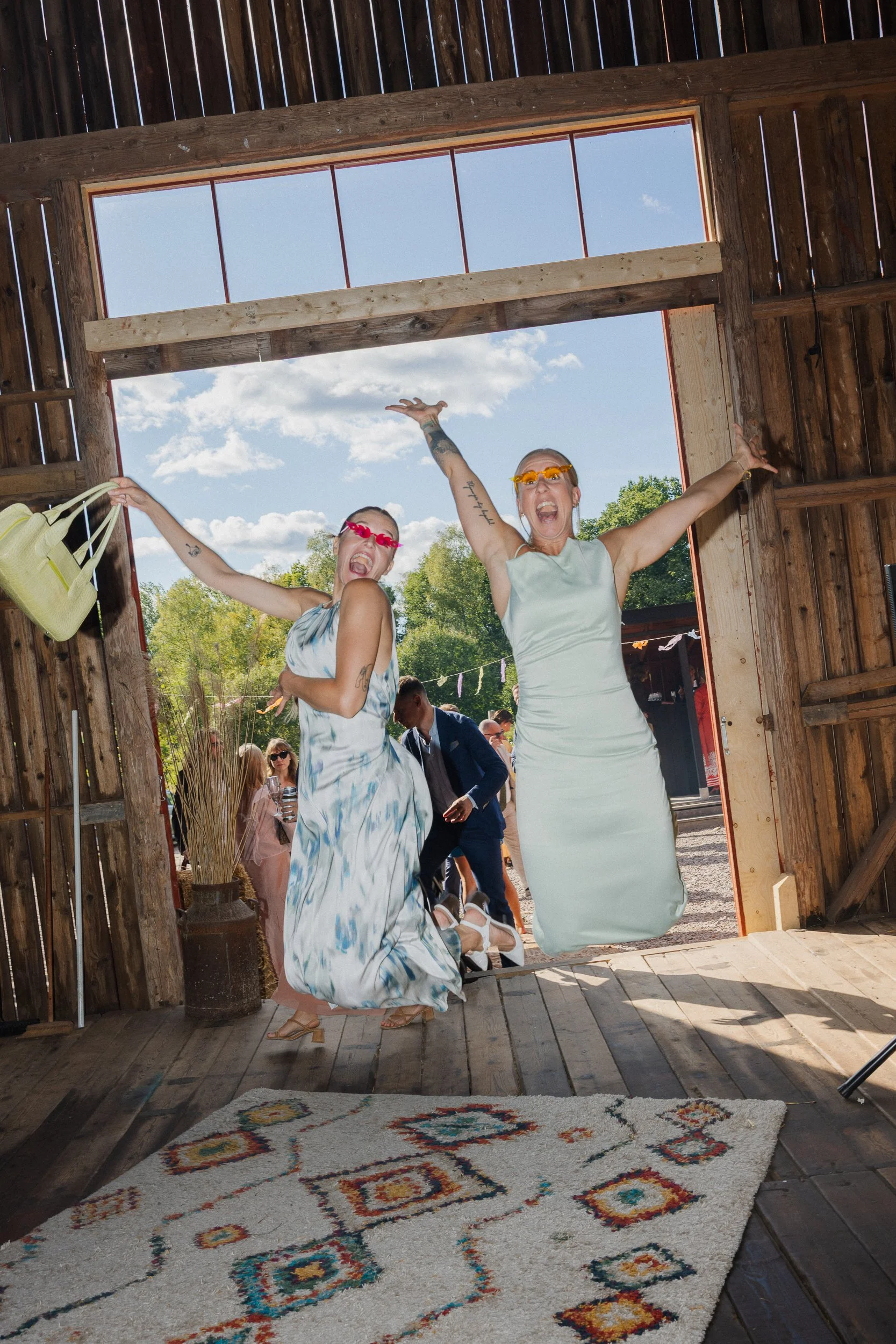 Två kvinnor som hoppar in i en lada under en fest, båda bär klänningar och solglasögon, medan gäster tittar på utomhus under en solig dag.  Av Sofia Wilhelmina bröllopsfotograf i Stockholm.