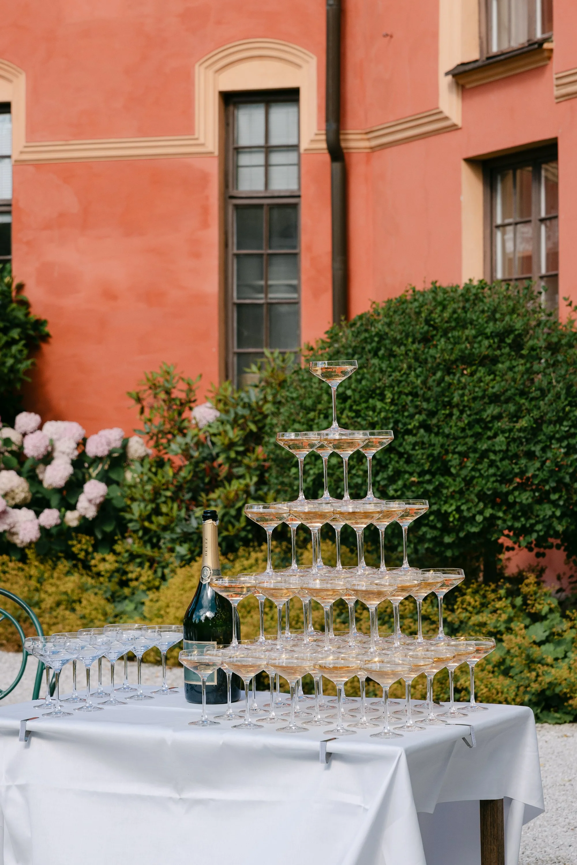 En förgylld champagnesamling på en vit duk täckt bord utomhus med ett rosa hus i bakgrunden.