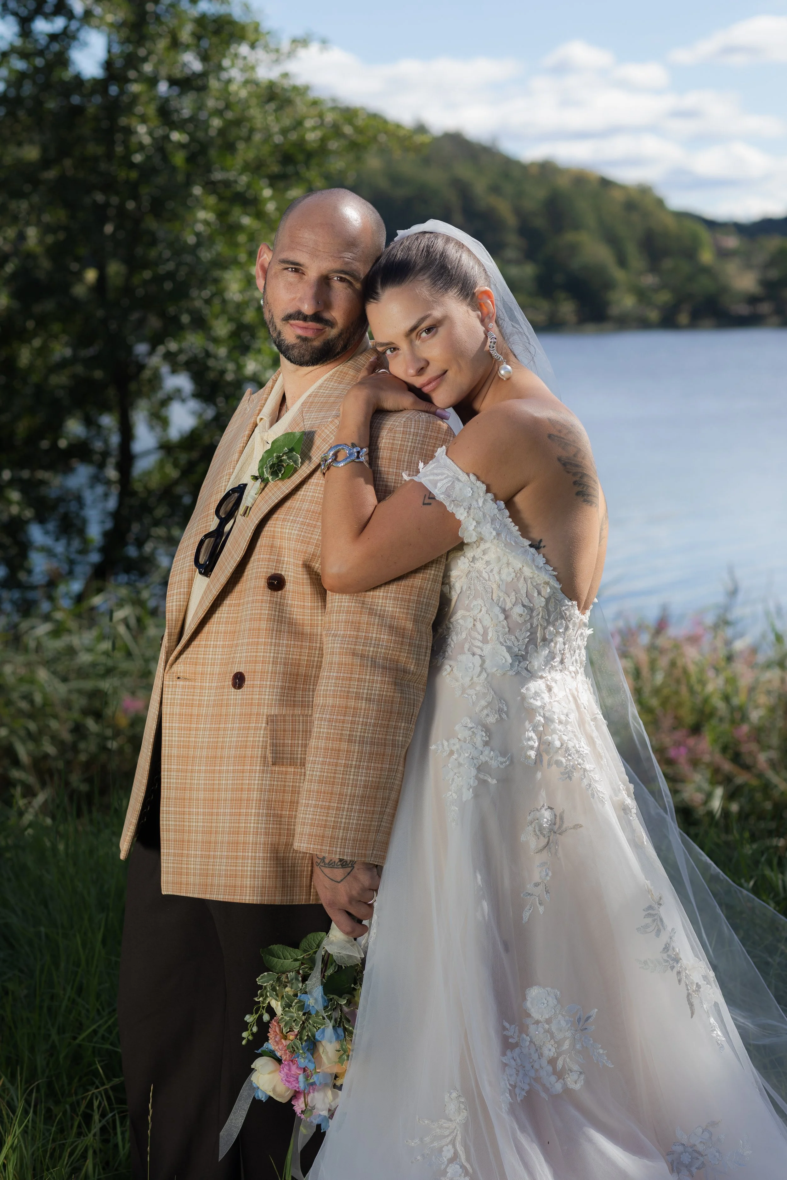 Ett brudpar står nära varandra utomhus vid en sjö, omgivna av träd och grönska, under en molnig himmel.