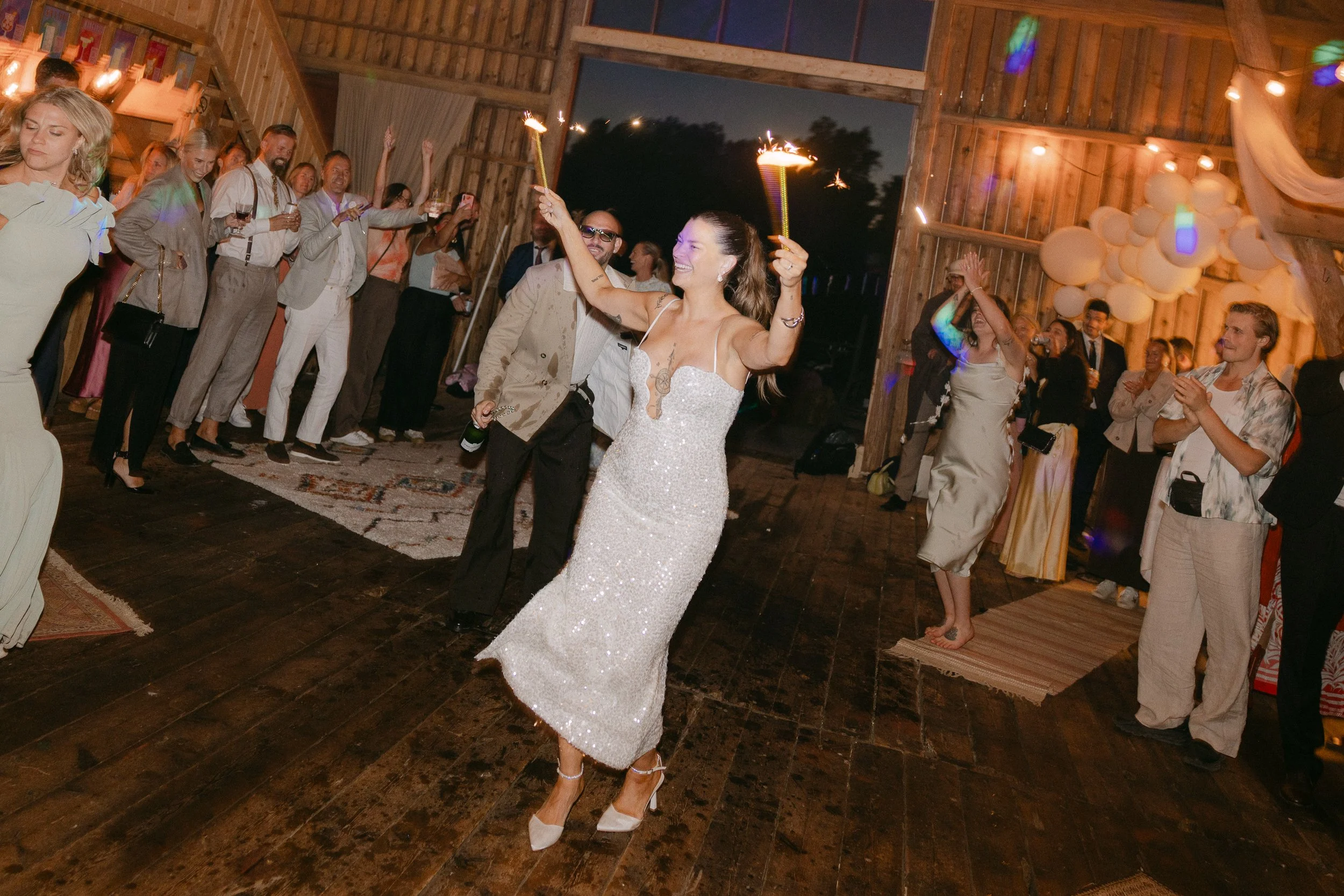 En brud i en vit, glittrande klänning dansar och leker med en tänd fackla under en fest. Runt omkring står gäster och tittar, många med drycker i handen, i ett rustikt trähus med varm belysning och dekorationer.