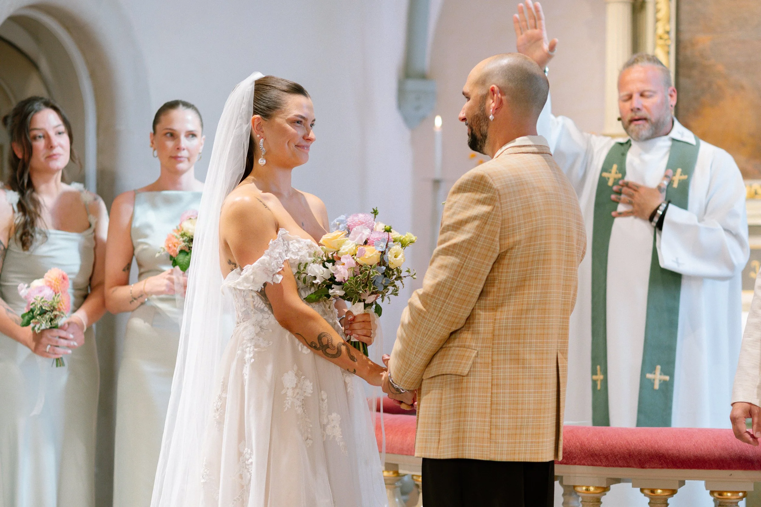 En vigsel i en kyrka där en kvinna i bröllopsklänning håller en bukett och tittar på sin blivande man i en ljusbrun kostym. En man i en kyrklig klädsel står bakom dem.  Av Sofia Wilhelmina bröllopsfotograf i Stockholm.