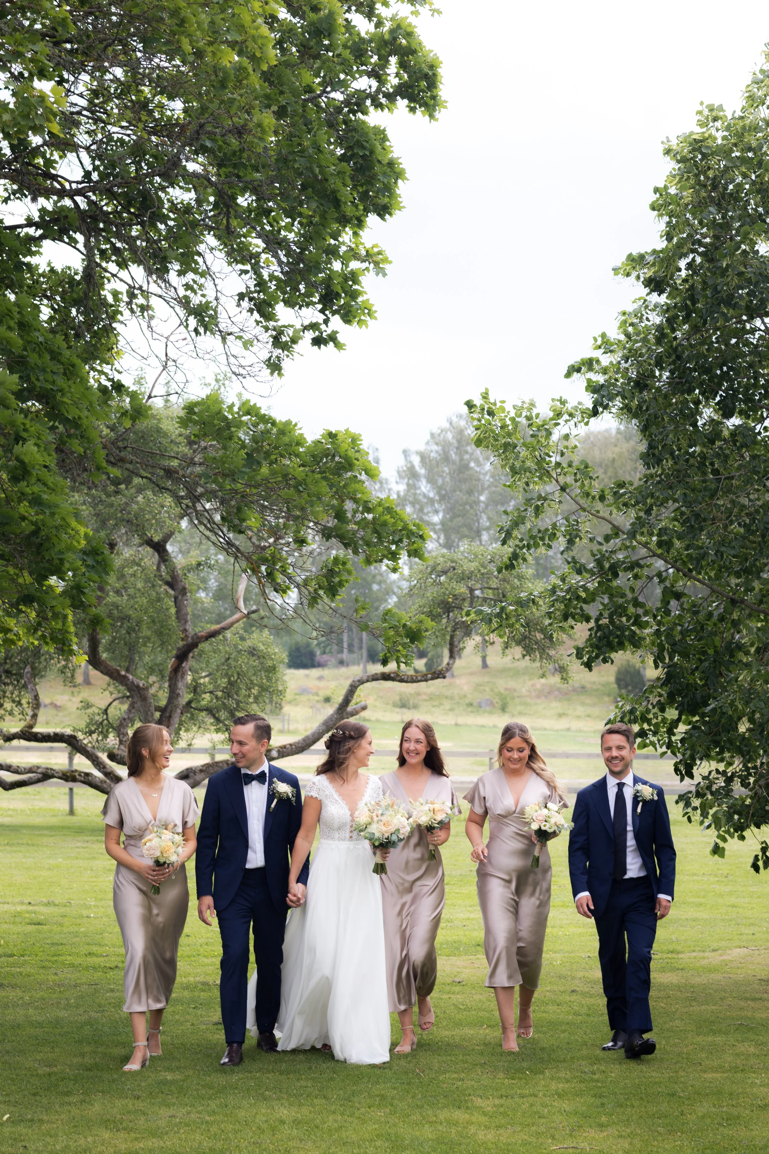 Bröllopspar och brudtärnor promenerar på gräset i en park med stora träd i bakgrunden.