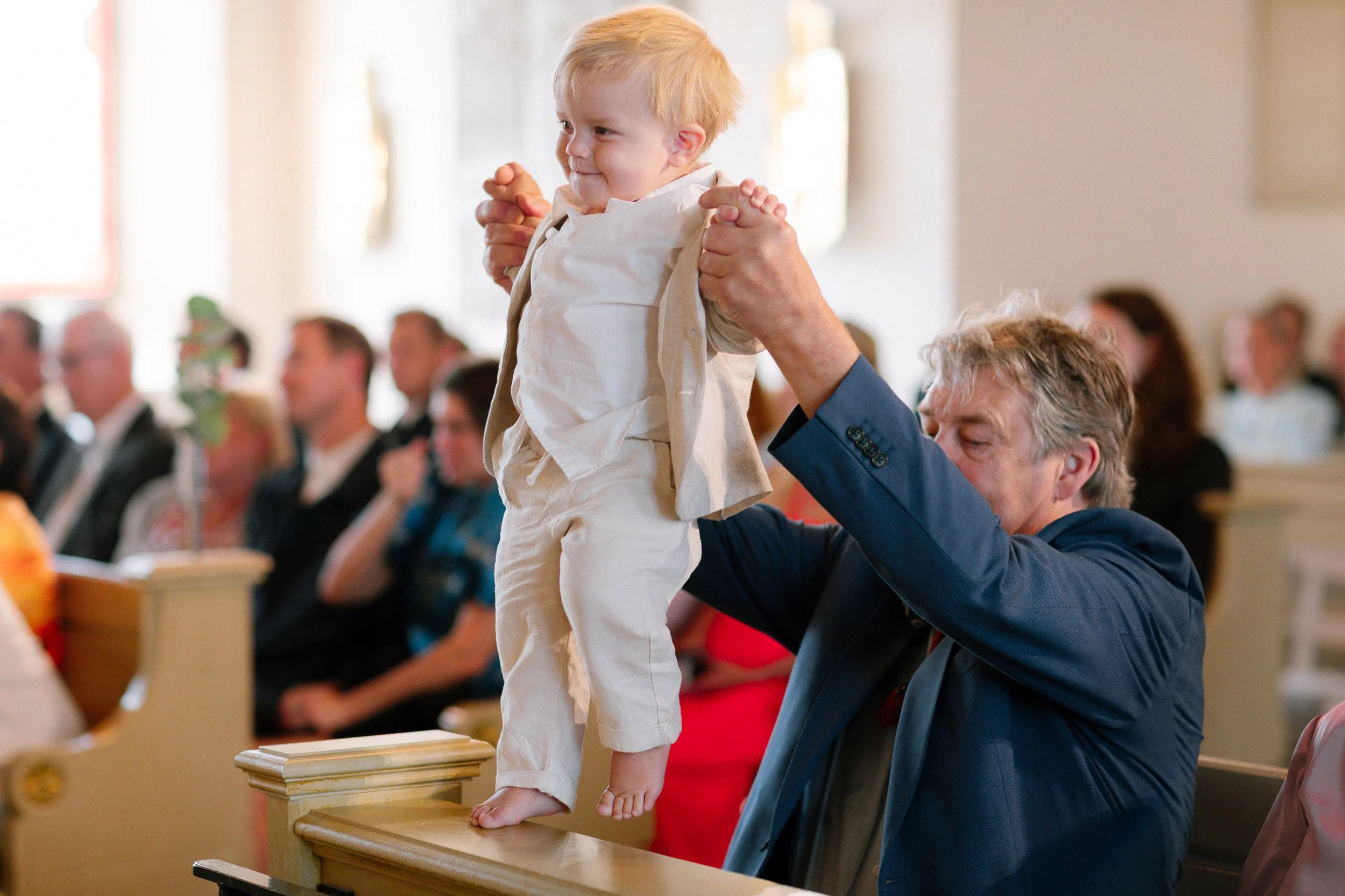 En glad liten pojke med blond hår som hålls uppe av en äldre man i en blå kostym under en ceremoni i kyrkan.