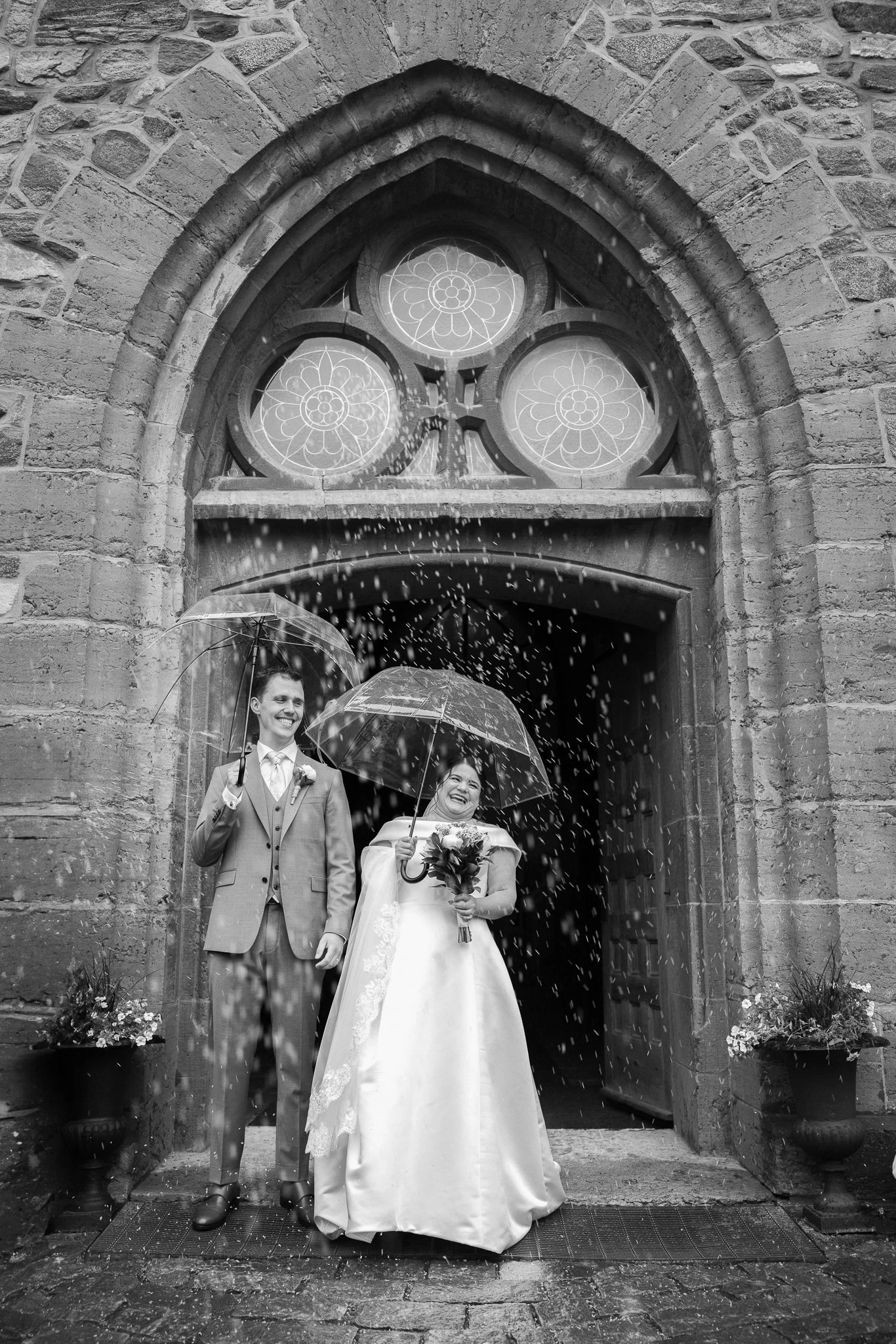 Två personer i bröllopskläder ler och står med paraplyer utanför en kyrka medan det regnar.