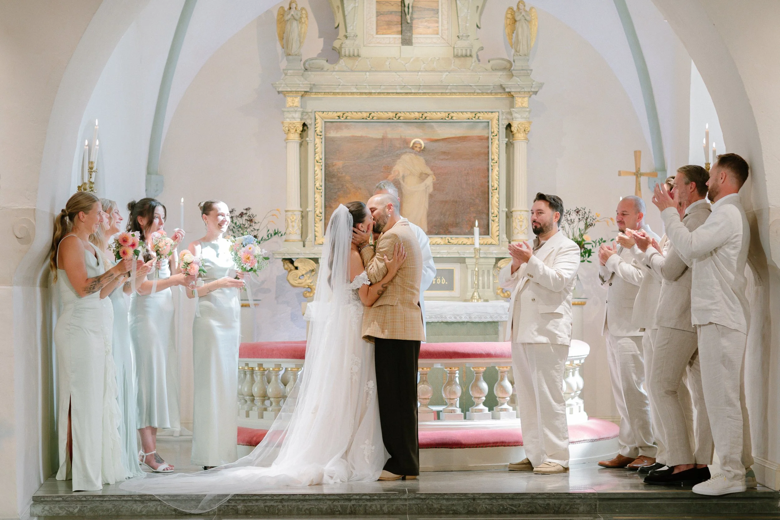 En brud och en brudgum kysser varandra i en kyrka under ett bröllop, omgiven av vitt klädda gäster som klappar och håller blombuketter.  Av Sofia Wilhelmina bröllopsfotograf i Stockholm.