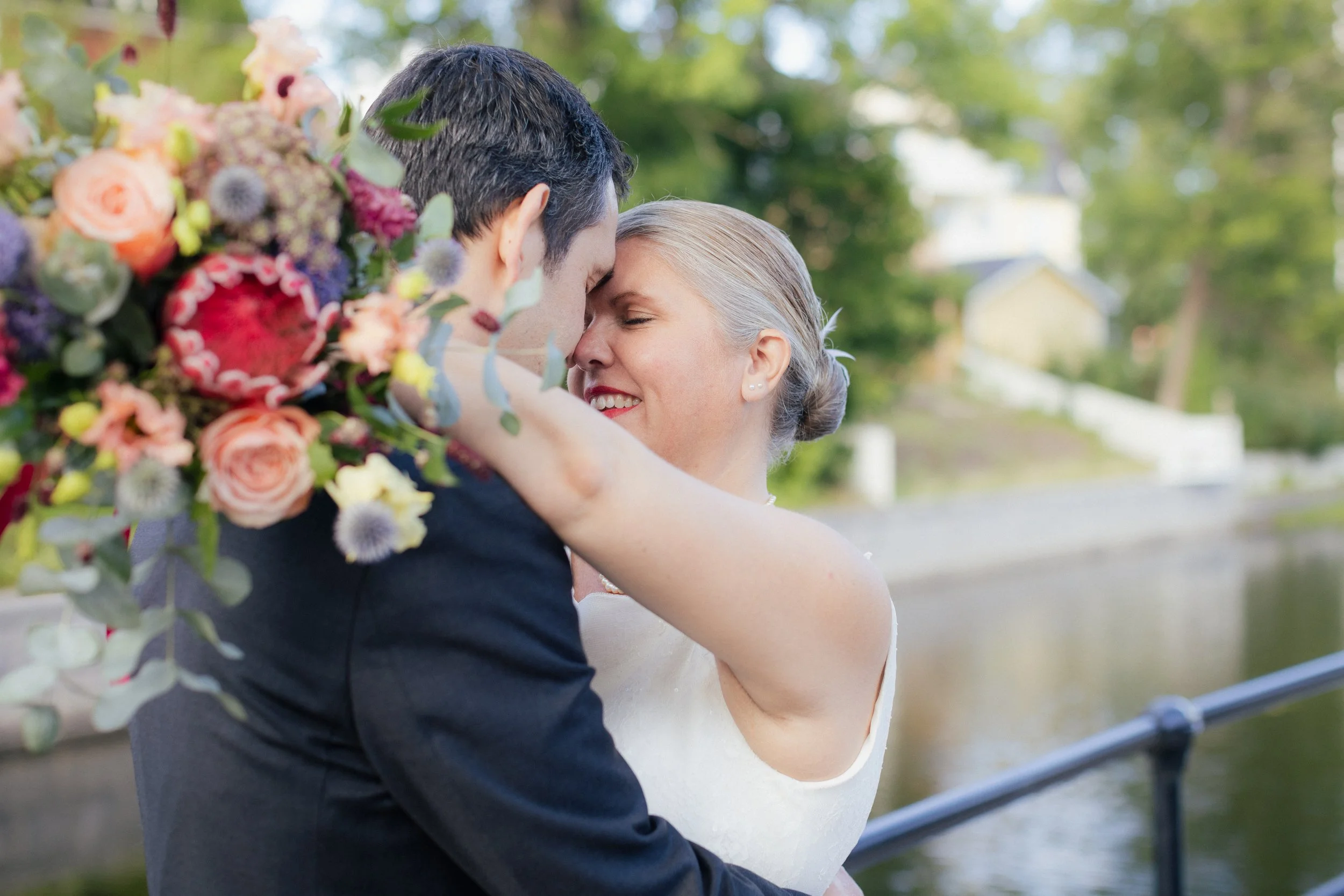 En lycklig brud och brudgumma omfamnar varandra nära en flod, med gröna träd och hus i bakgrunden.