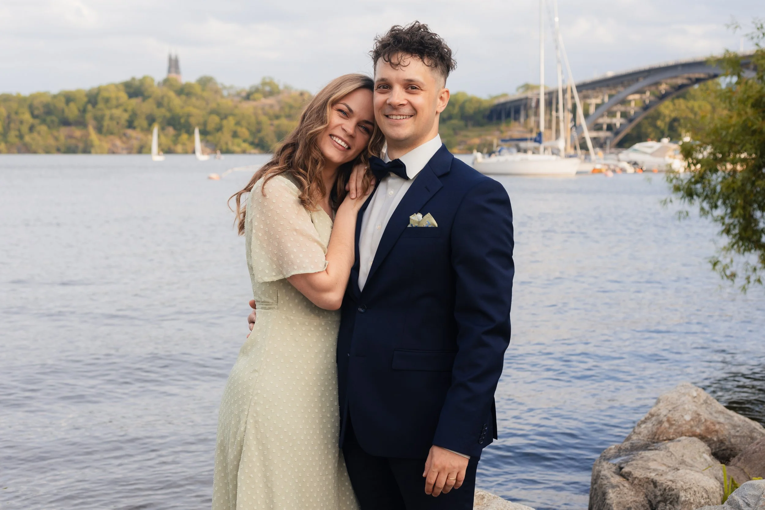 En glad man och en kvinna poserar vid vattnet, klädda i formella kläder, med båtar och en bro i bakgrunden.