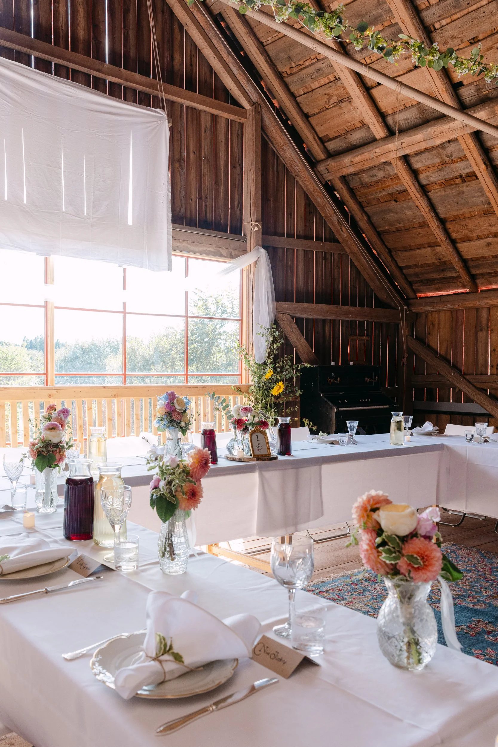 Varmt inbjudande middagsrum med högt tak och träväggar. Bord är dukade för en fest eller bröllopsmiddag med blommor, glas, servetter, och ljus. Ett stort fönster släpper in mycket dagsljus och visar gröna träd utanför.