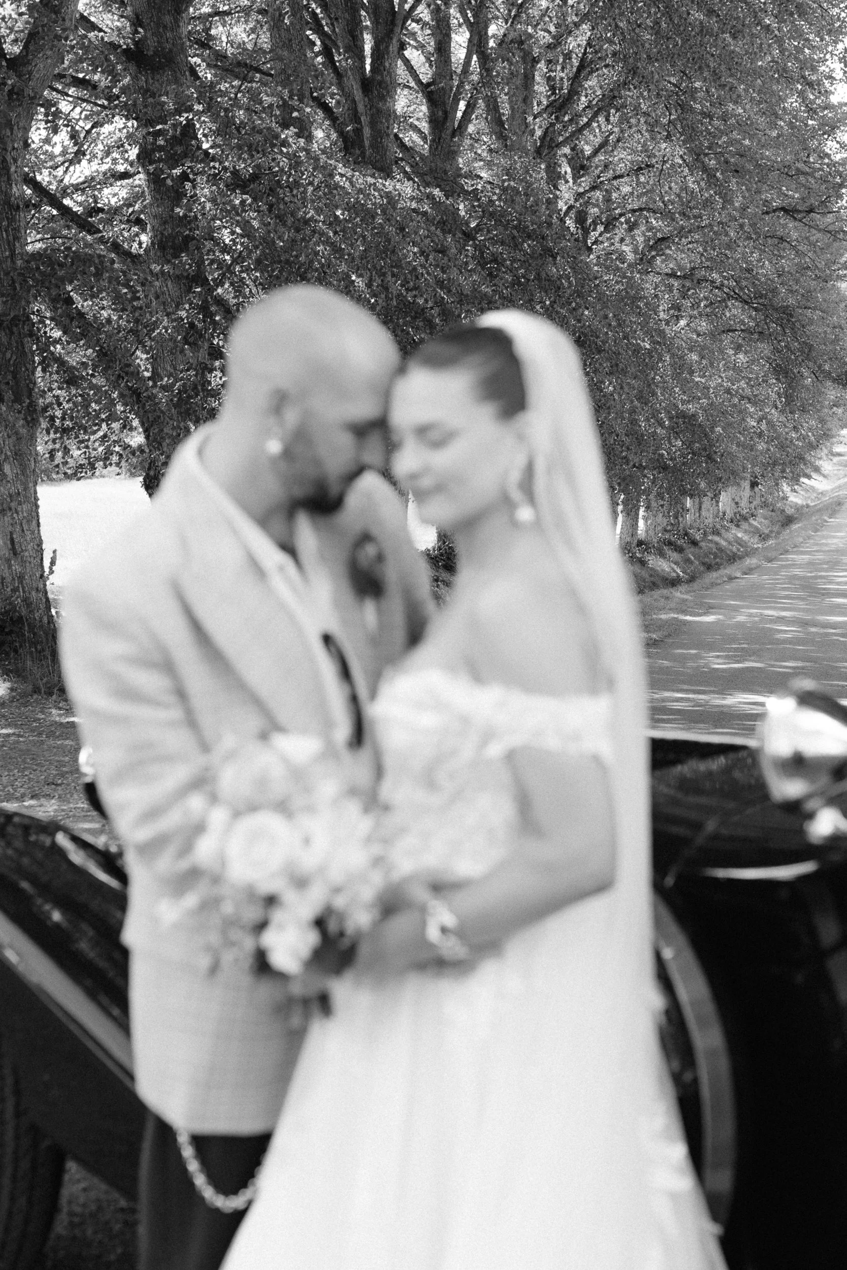 Två människor på bröllopsdag, står nära varandra med ansikten nära, framför en linje av träd längs en väg, i svartvitt.  Av Sofia Wilhelmina bröllopsfotograf i Stockholm.