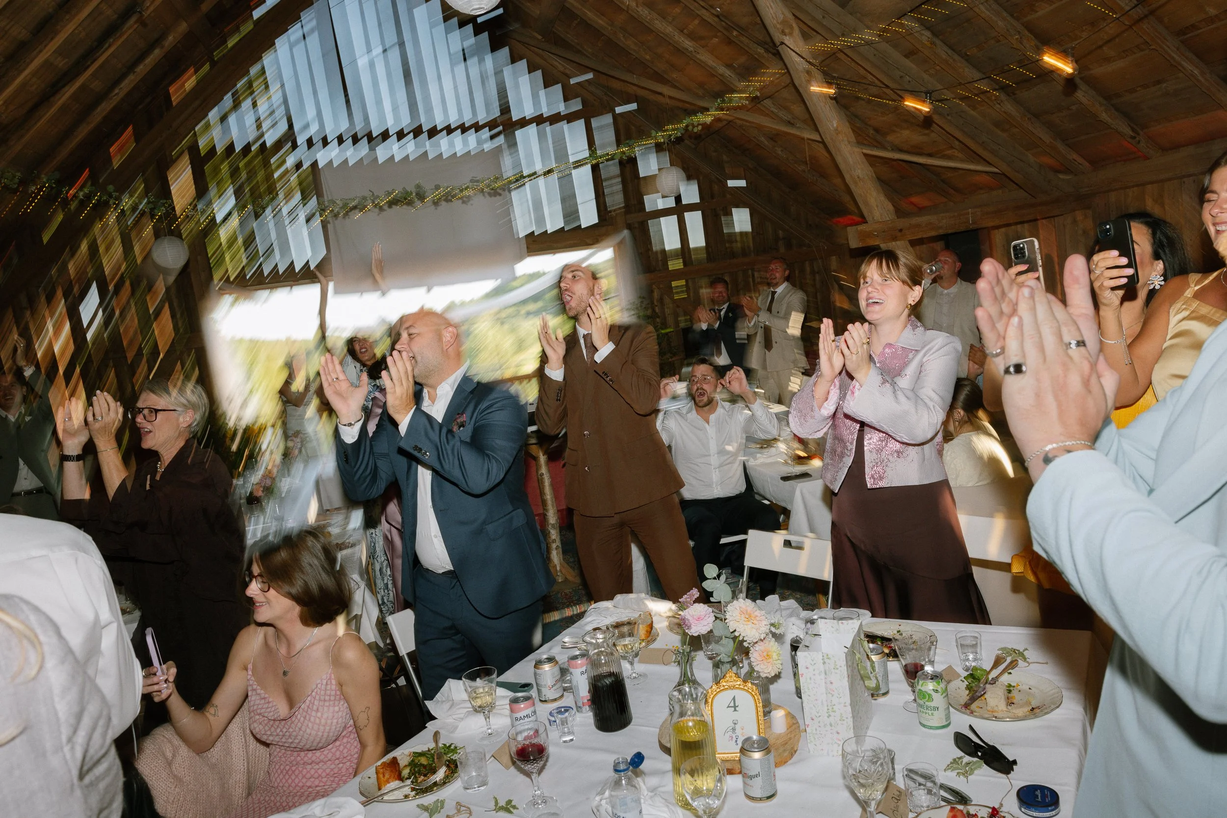 Folk som står och klappar på ett festbord i ett träloft, några som skrattar och tar bilder, samt person i bröllopskläder som dansar och firar.
