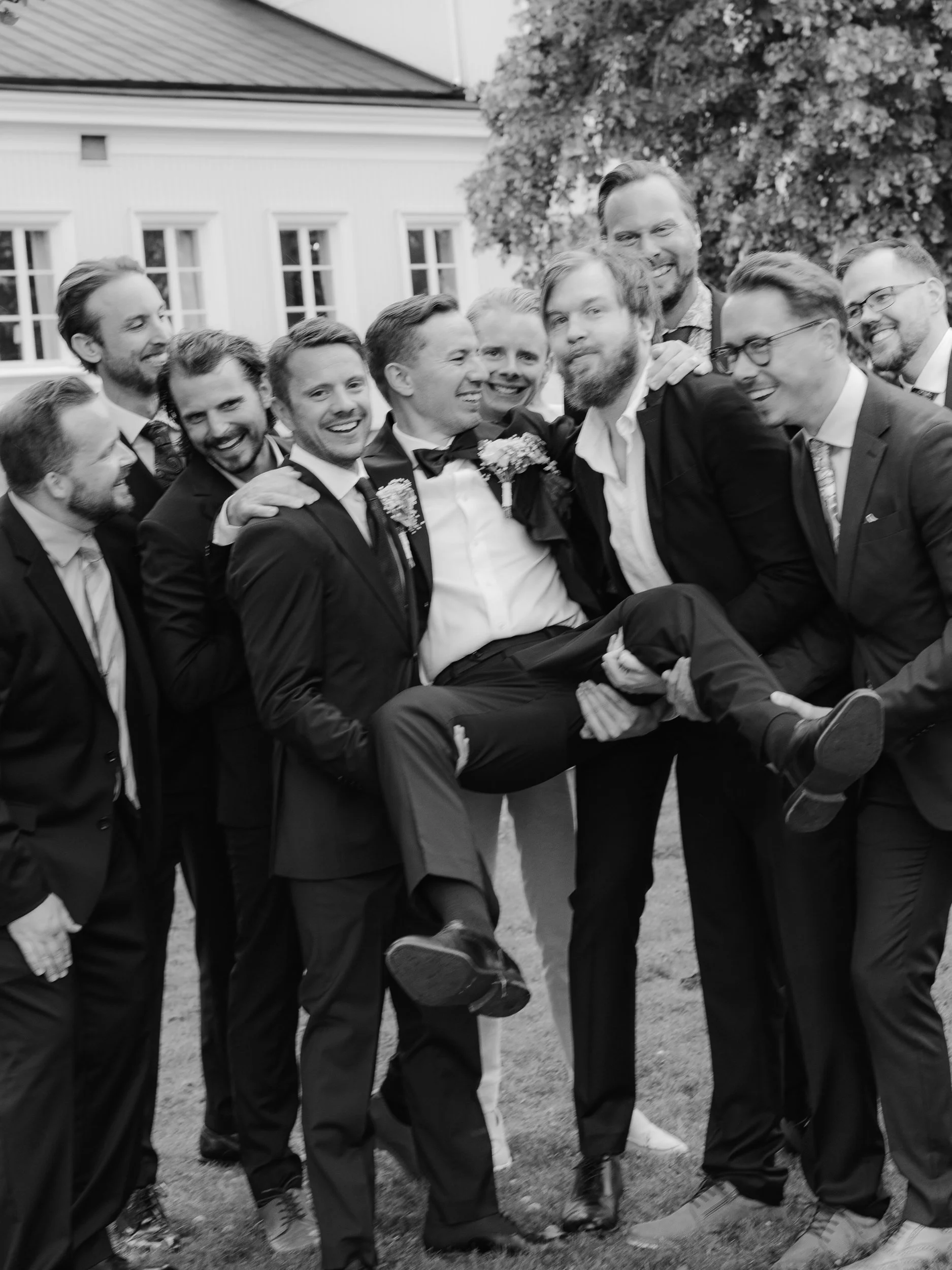 En grupp män i kostymer som firar, en människa blir buren av andra i gruppen, utomhus med träd och ett hus i bakgrunden.