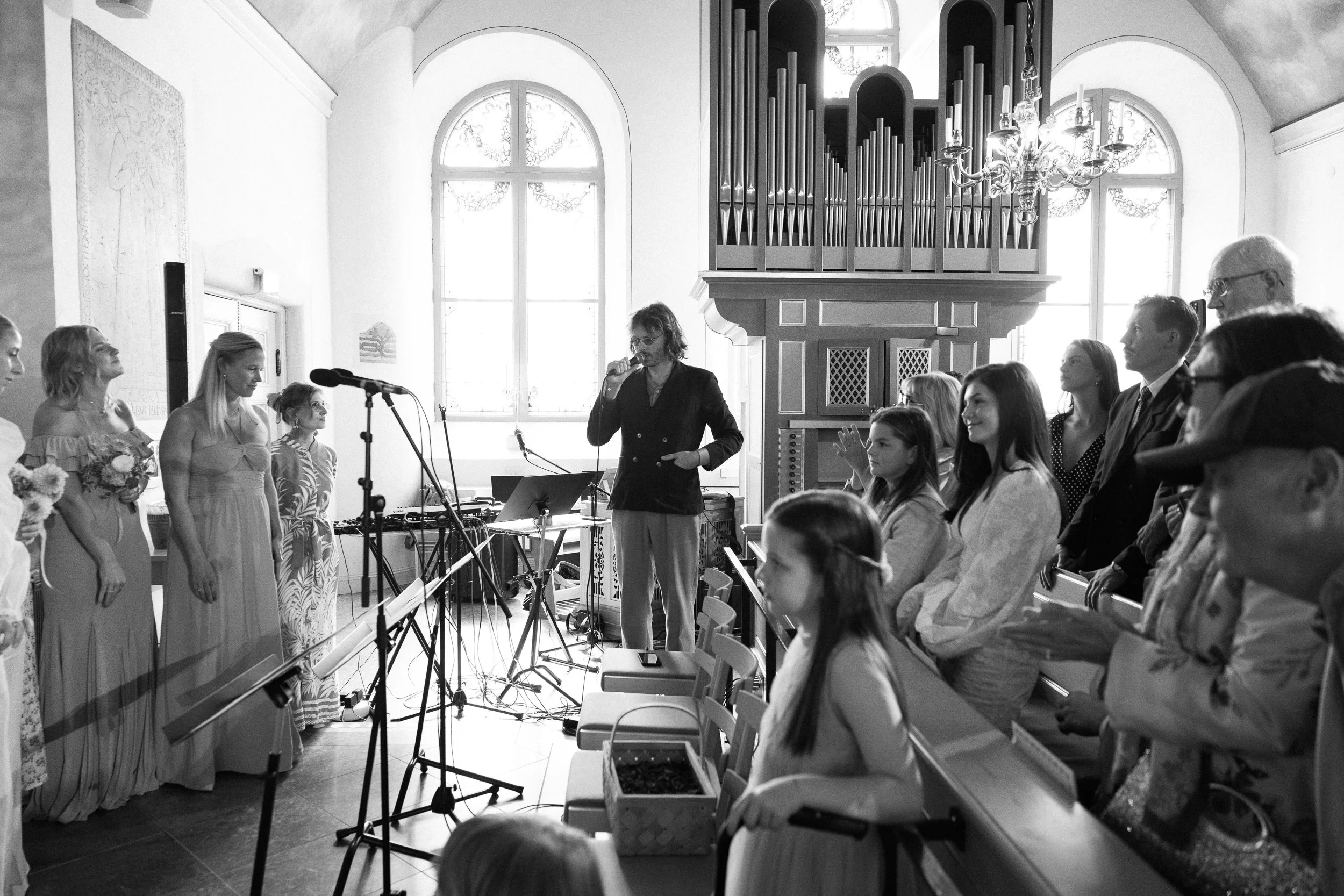 Gäster i kyrka vid musikframträdande, kvinnor i vackra klänningar och män i kostymer, ungar och äldre tittar på, några personer håller i blommor, bilder av orgel och stora fönster i bakgrunden.