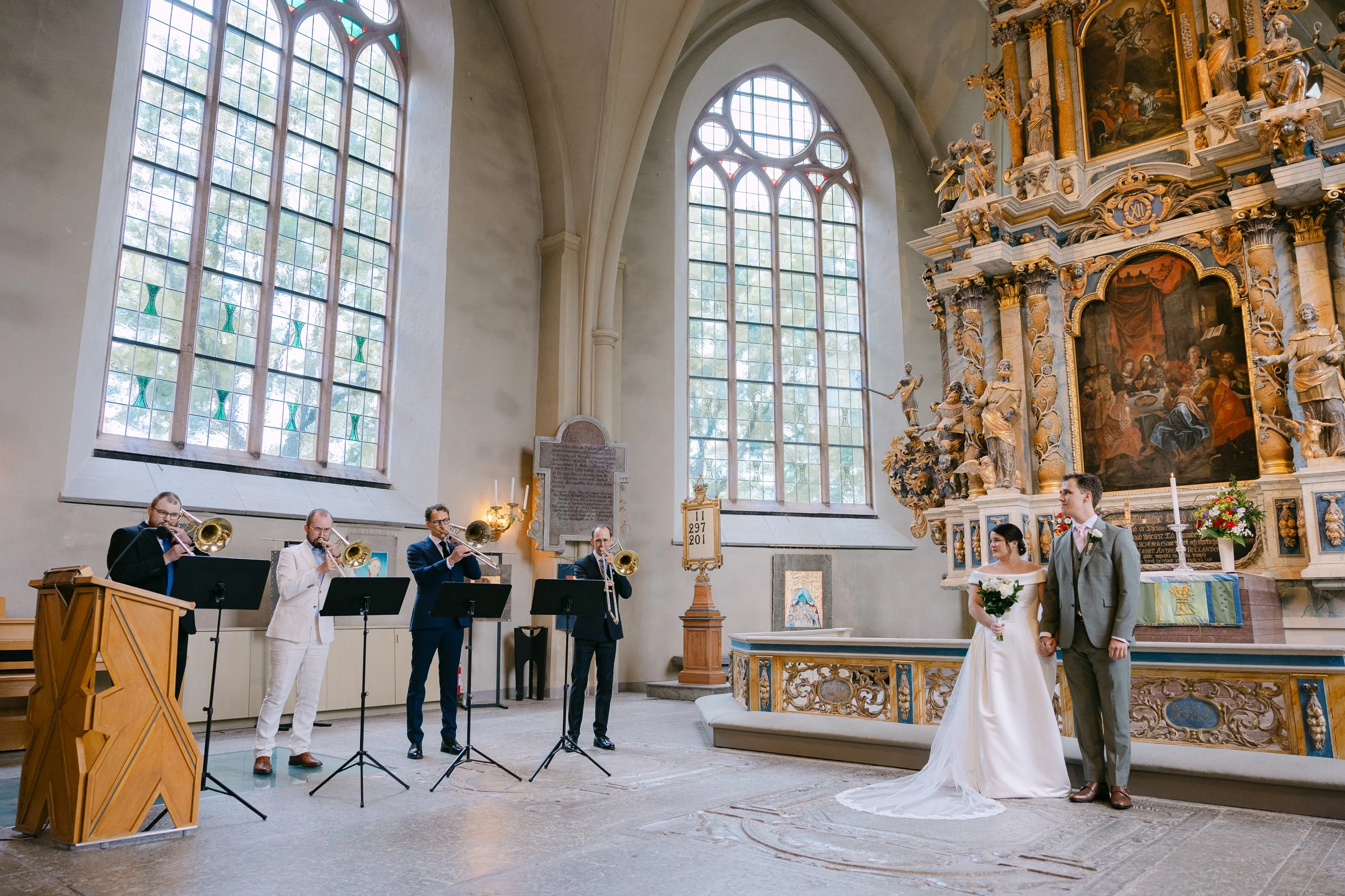 Brudpar som står framför altaret i en kyrka under ett bröllop, med ett bröllopsband. Musiker spelar i bakgrunden, stora fönster släpper in ljus.