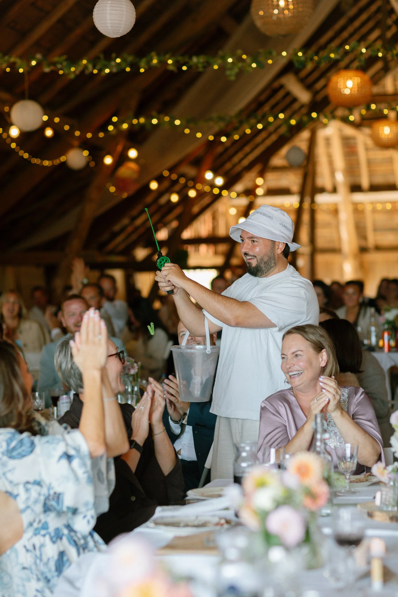 En man i vit klädsel och hatt under en ceremoni, som sprutar champagne på en fest med många glada gäster i en träbod med juldekorationer och ljusslingor.  Av Sofia Wilhelmina bröllopsfotograf i Stockholm.