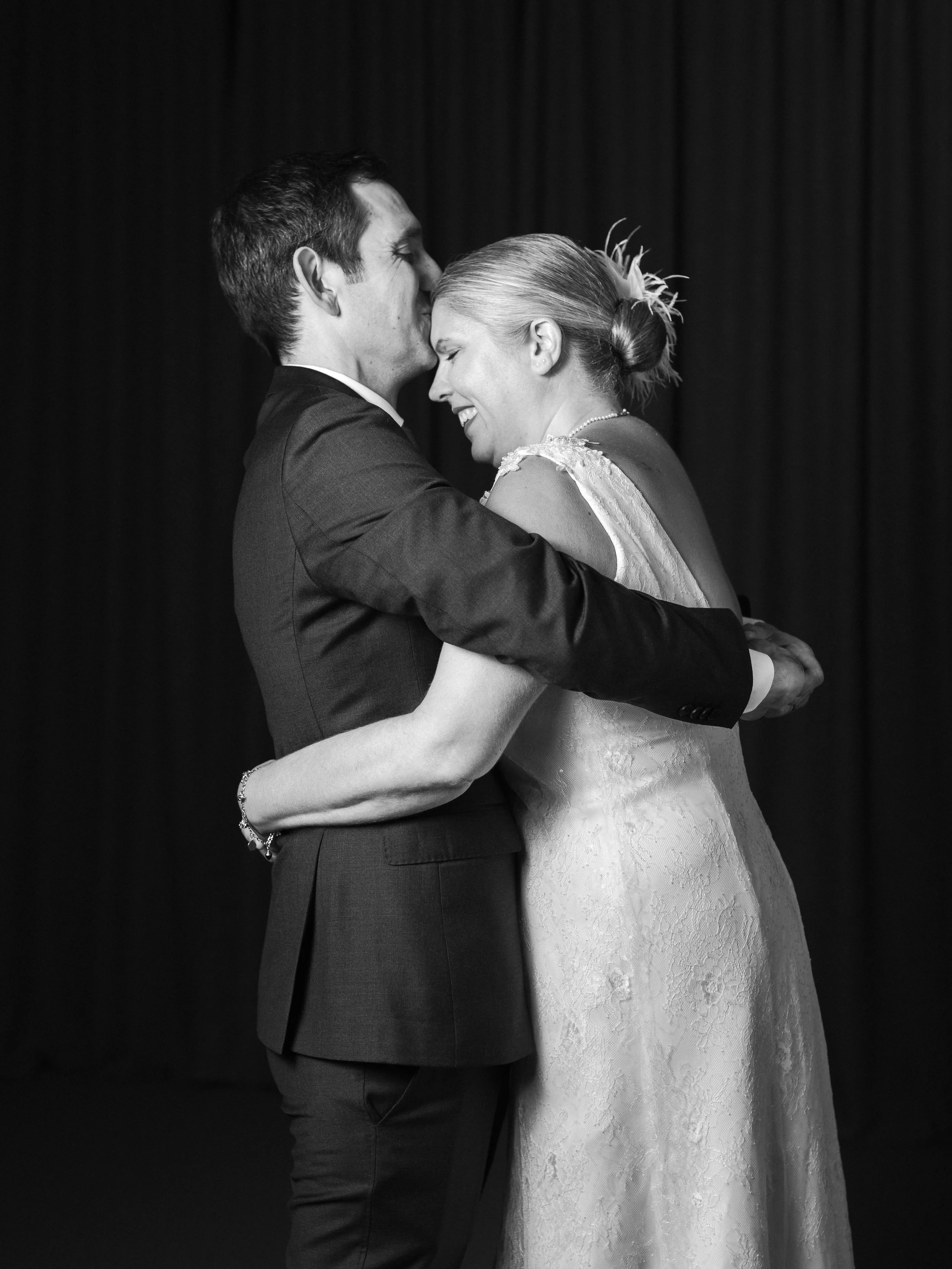 Svartvitt foto av ett brudpar som kysser och håller om varandra, med mörk bakgrund.