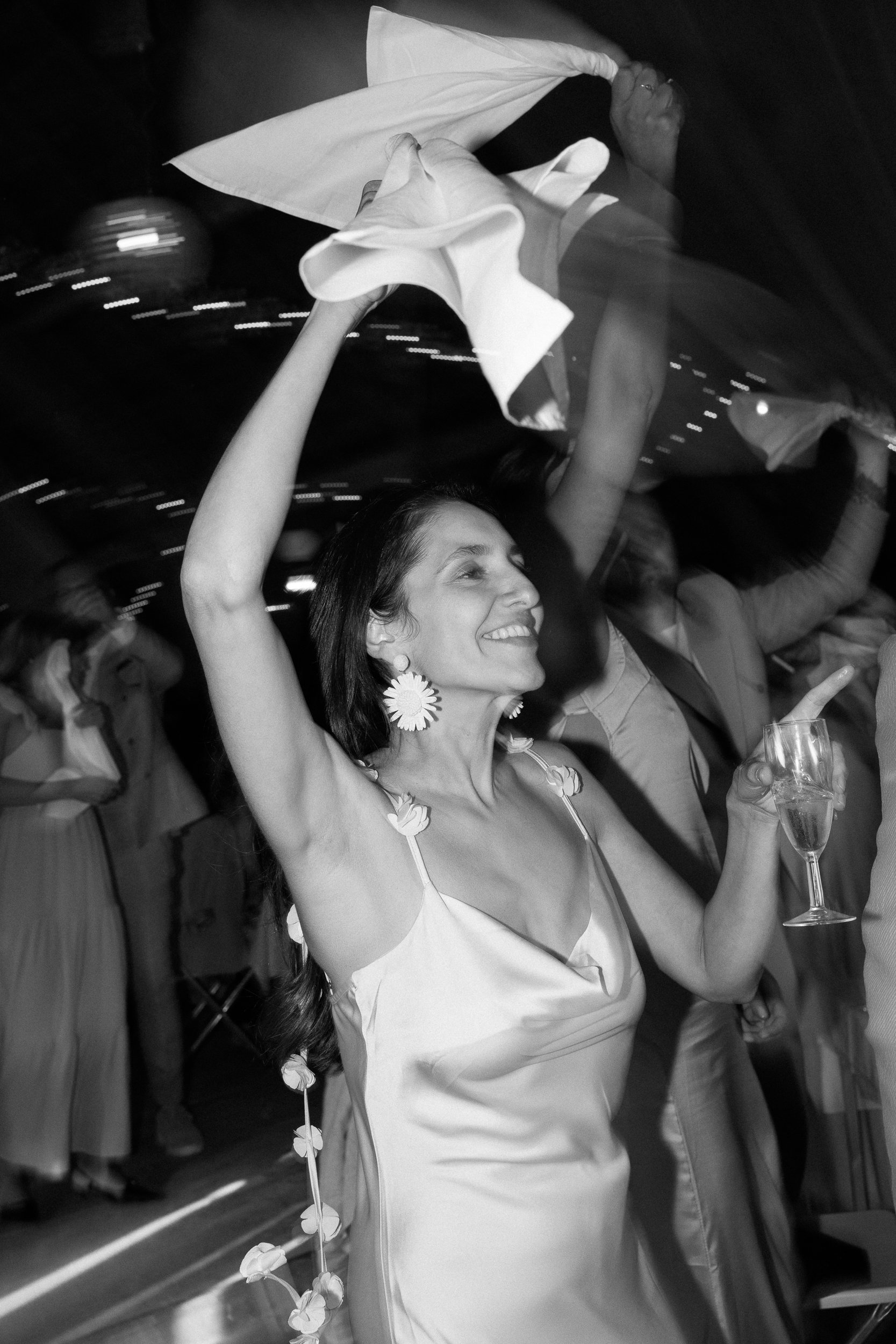 En kvinna som ler och dansar iklädd en ljus klänning med blomsterdetaljer, hon håller ett glas vin i handen och har stora blomsterörhängen. Hon tycks vara glad och firar, omgiven av andra människor i en festlig atmosfär.  Av Sofia Wilhelmina bröllops