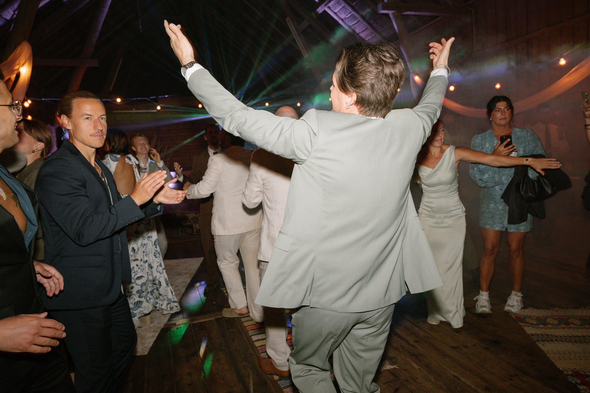 Flera människor dansar på ett dansgolv med disco- och ljuseffekter, en man i ljus kostym står med armarna utsträckta framför andra.  Av Sofia Wilhelmina bröllopsfotograf i Stockholm.