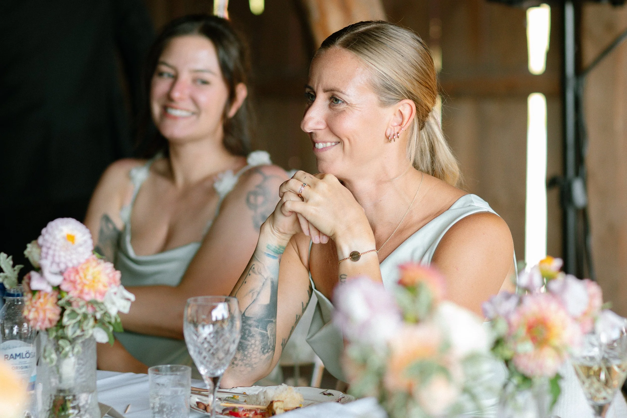 Två kvinnor sitter vid ett bord, en av dem ler och den andra ser glad ut, och de är klädda i ljusa festkläder, med blommor som dekorationer på bordet, i en rustik träbyggnad.