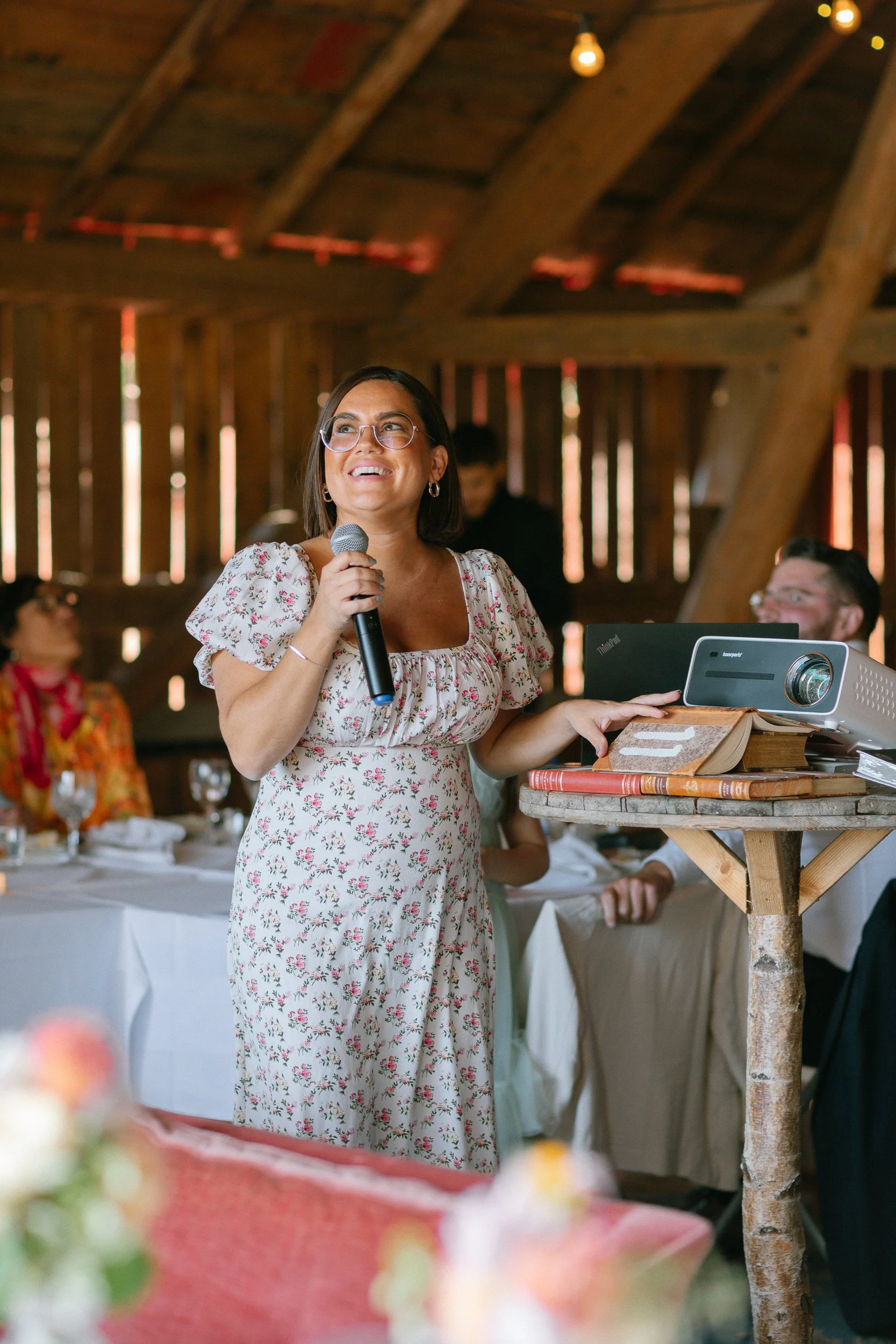 Kvinna som håller en mikrofon och pratar på ett bröllop eller fest, med flera personer och dukade bord i bakgrunden i ett trähus.