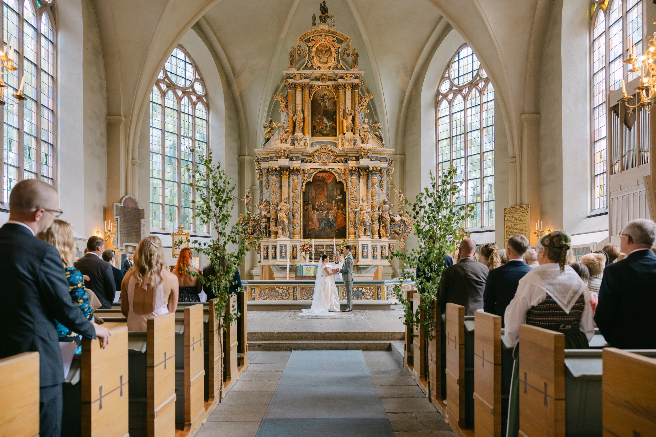 Ett brudpar står framför prästen i en kyrka under deras bröllopsceremoni, omgivna av gäster som sitter på bänkar. Kyrkan har stora fönster, höga valv och ett rikt dekorerat altarskåp i bakgrunden.