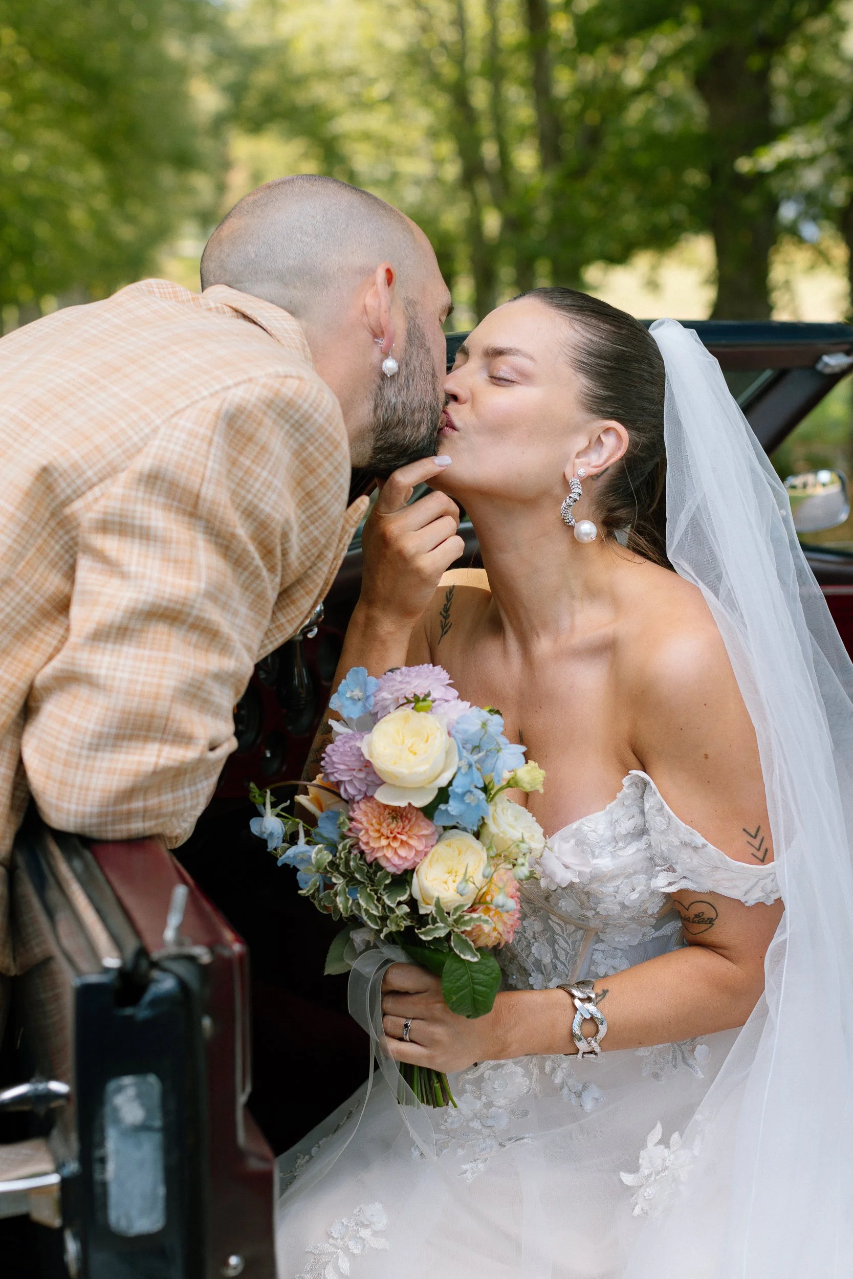 En brud och en man kysser varandra utomhus, hon bär en vit brudklänning med blommönster, stor vit slöja och stora örhängen, hon håller en bukett med färgglada blommor.  Av Sofia Wilhelmina bröllopsfotograf i Stockholm.
