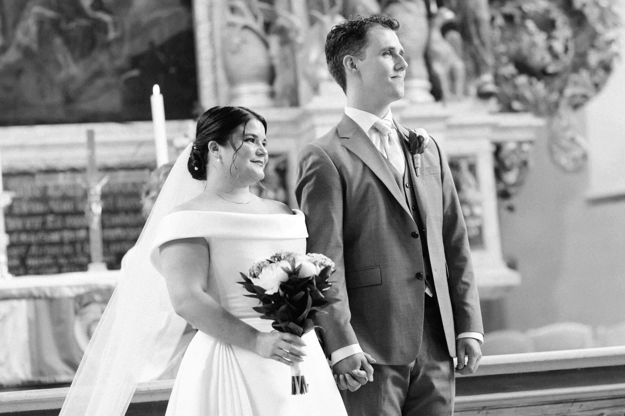 Ett brud- och brudgumpar som håller varandras händer under ett bröllop i en kyrka, med brudens bouquet i handen, i svartvitt.