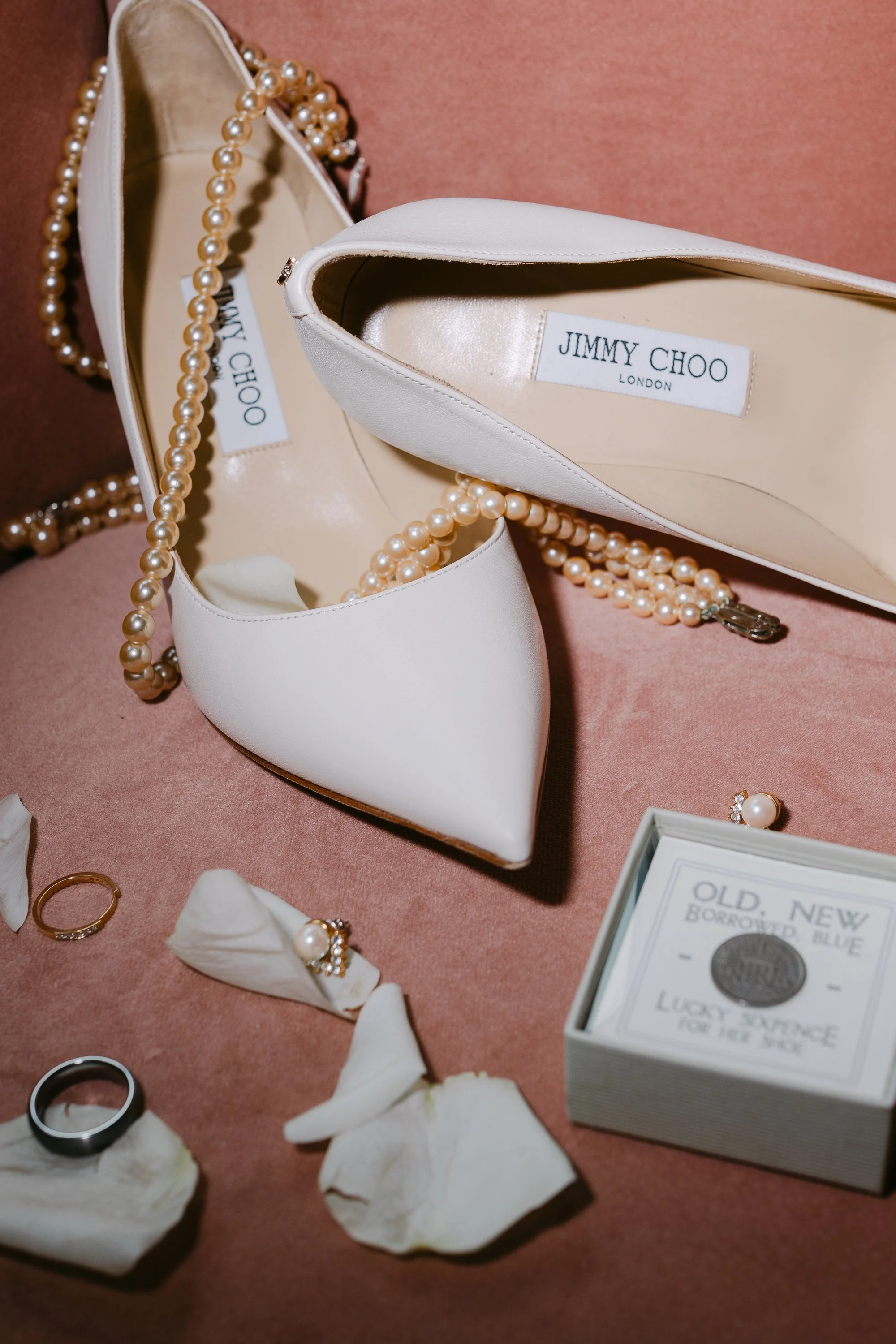 En vit Jimmy Choo damsko och ett pärlarmband, ett par örhängen med pärla och diamant, ringen, ett par svarta och vita ringar, ett ask med smycken och några blomblad på ett rosa tyg.