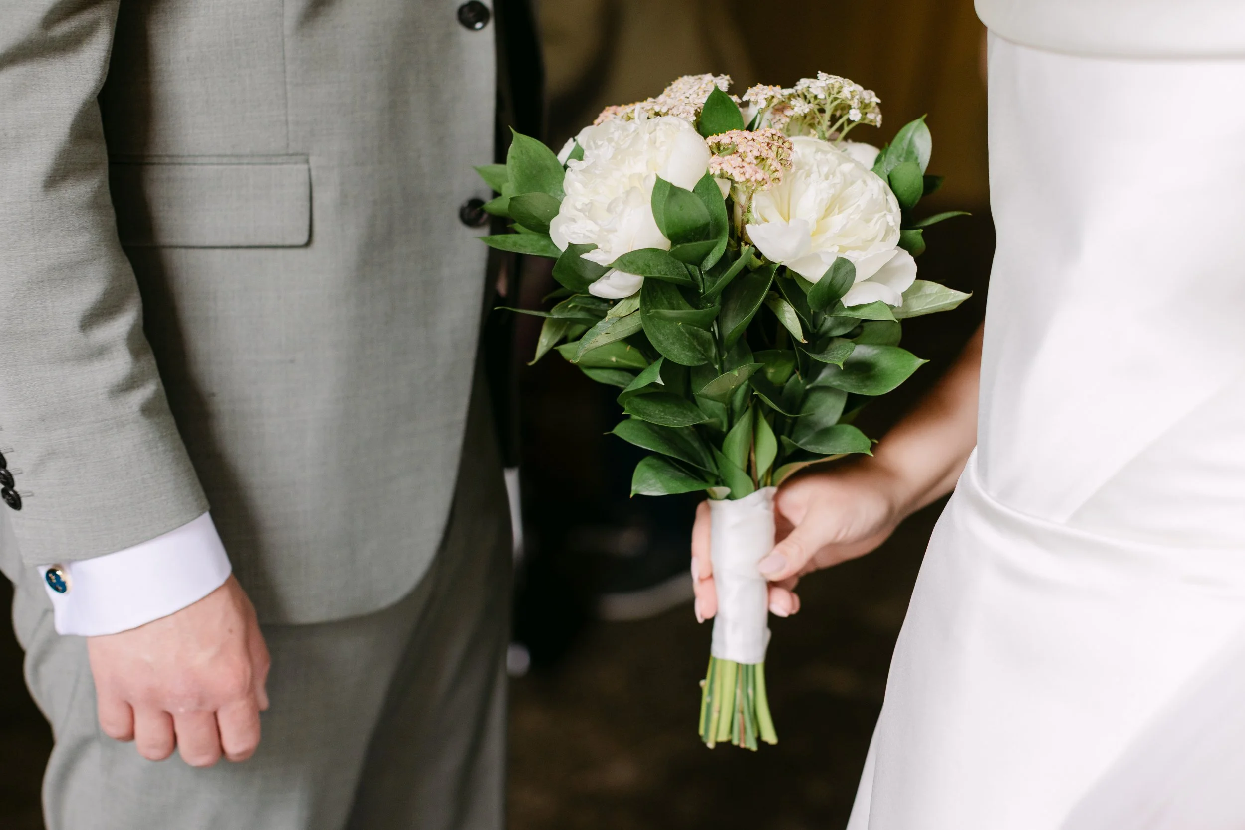 Två personer, en man och en kvinna, håller i en blomsterbukett med vita blommor och grönt blad. Mannens händer och del av hans ljusgrå kostym är synliga, kvinnans vita ärmlösa klänning är också delvis synlig.