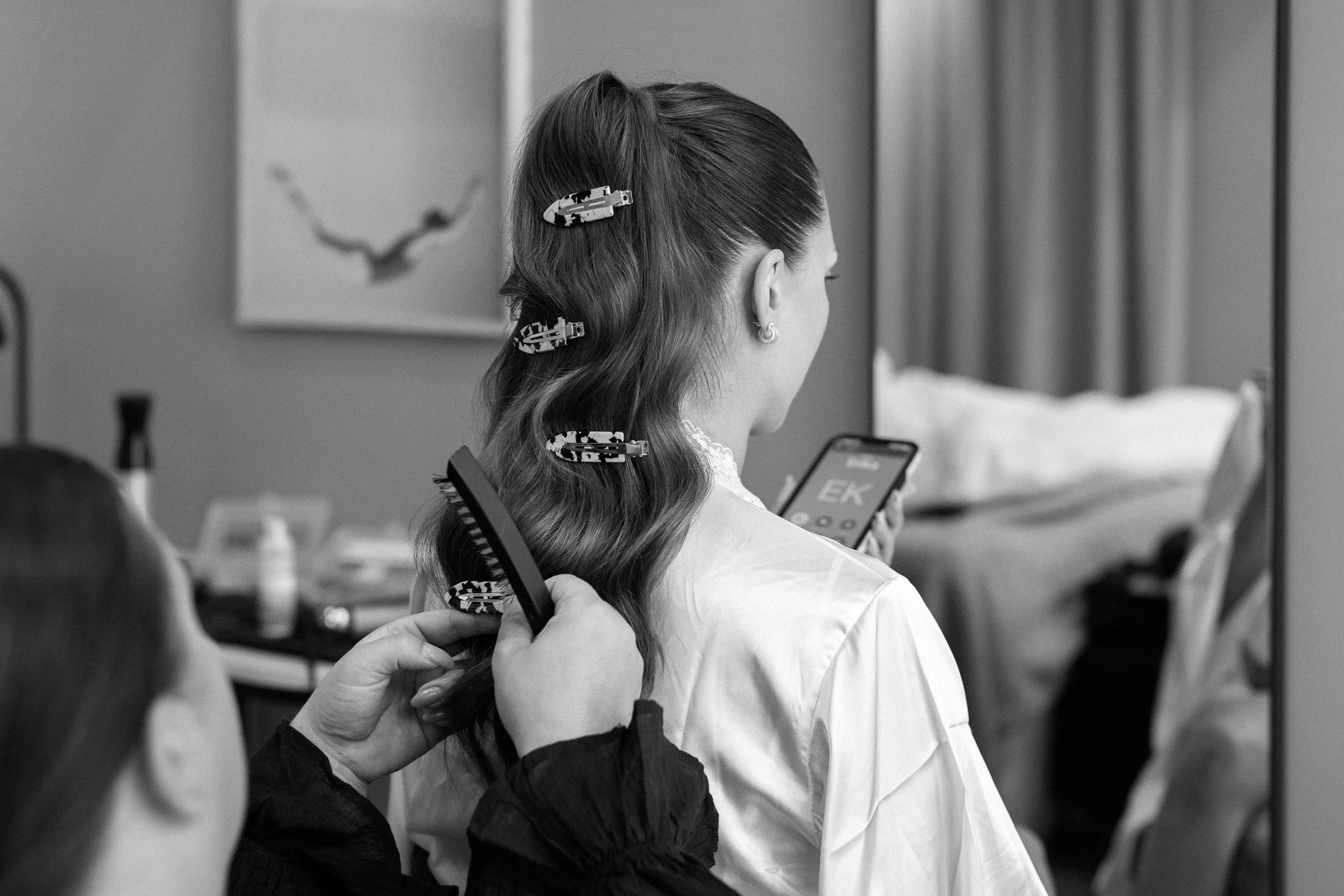 En kvinna som väntar på att bli stylad, någon ägnar sig åt hårstyling med klämma och borste, i ett inomhusrum.  Av Sofia Wilhelmina bröllopsfotograf i Stockholm.