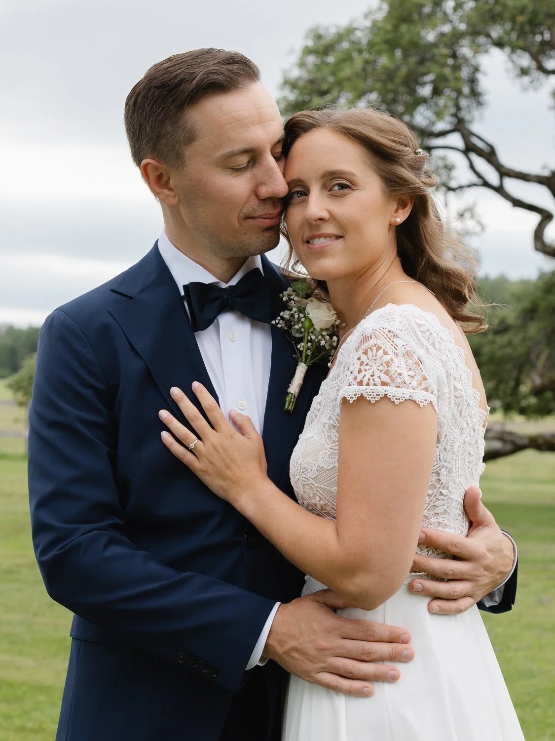 Ett brudpar omfamnar varandra utomhus, mannen i mörkblå kostym och kvinna i vit brudklänning, med träd och gräs i bakgrunden.