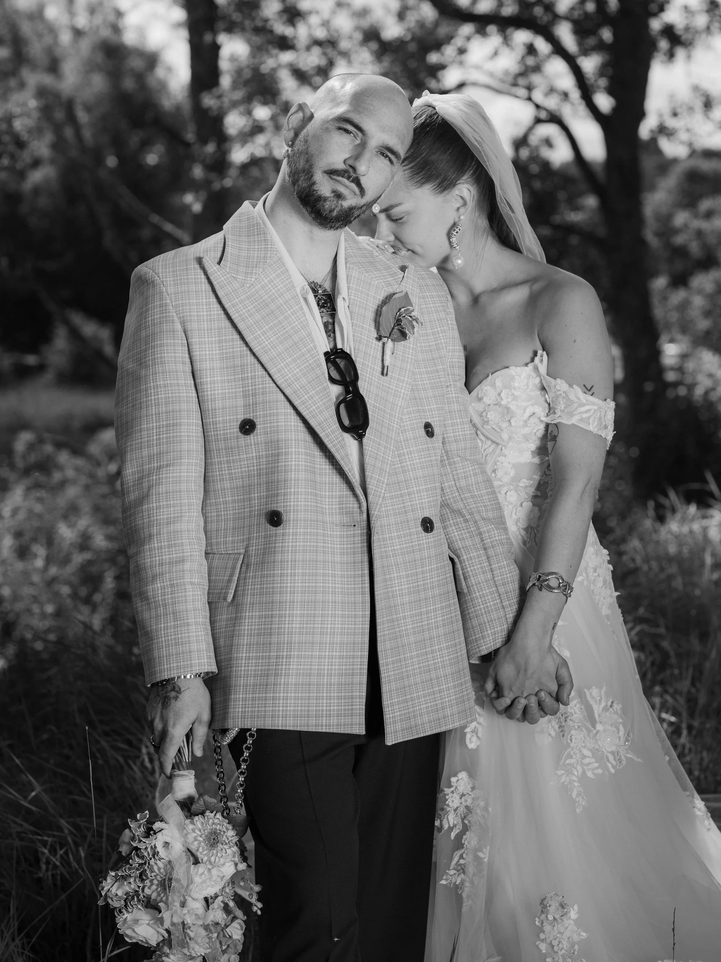 Ett brudpar som håller handen, hon vilar sitt huvud mot hans axel på en grön, utomhusbakgrund. Editorial look, båda är allvarliga och har ovanliga bröllopskläder.