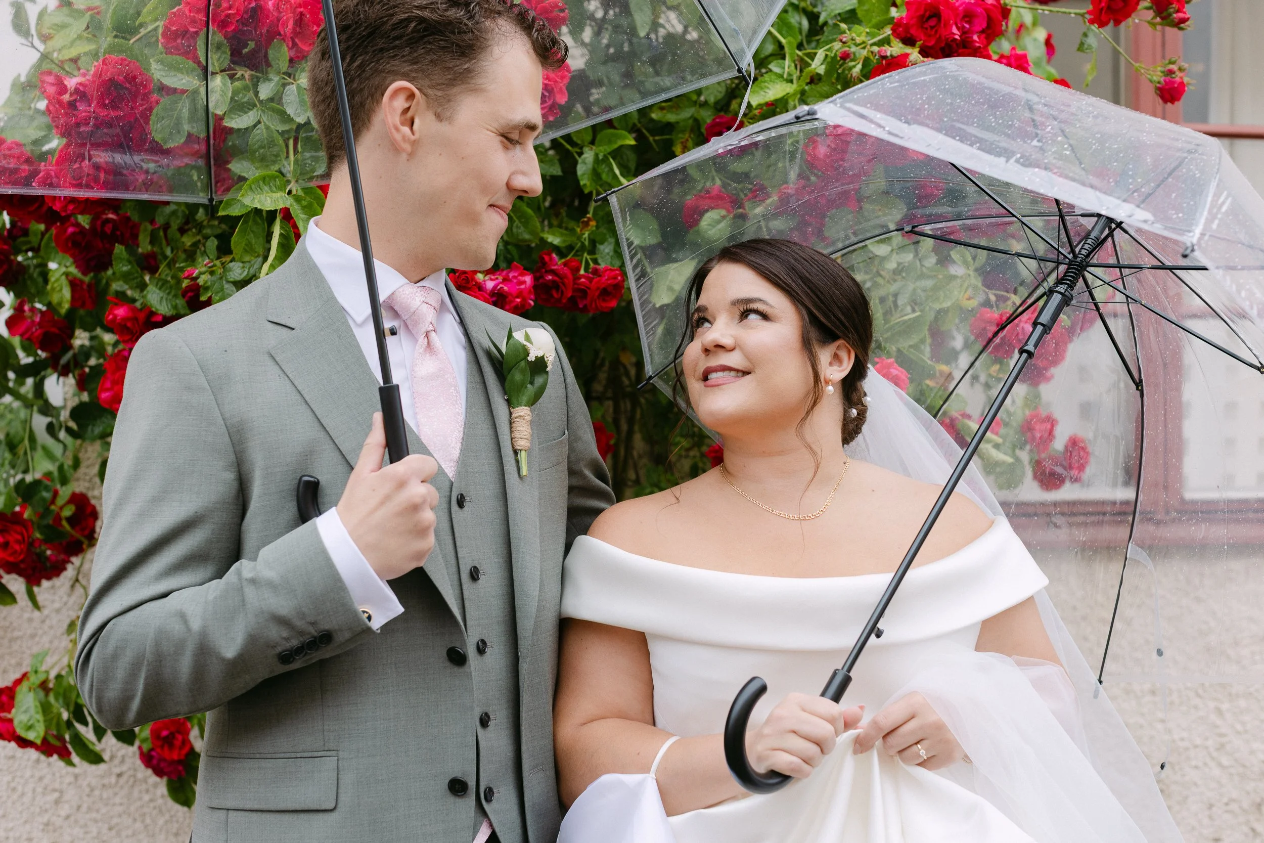 En brud och en brudgum står under ett transparent paraply utomhus, omgiven av röda blommor, och ler mot varandra i regnet.